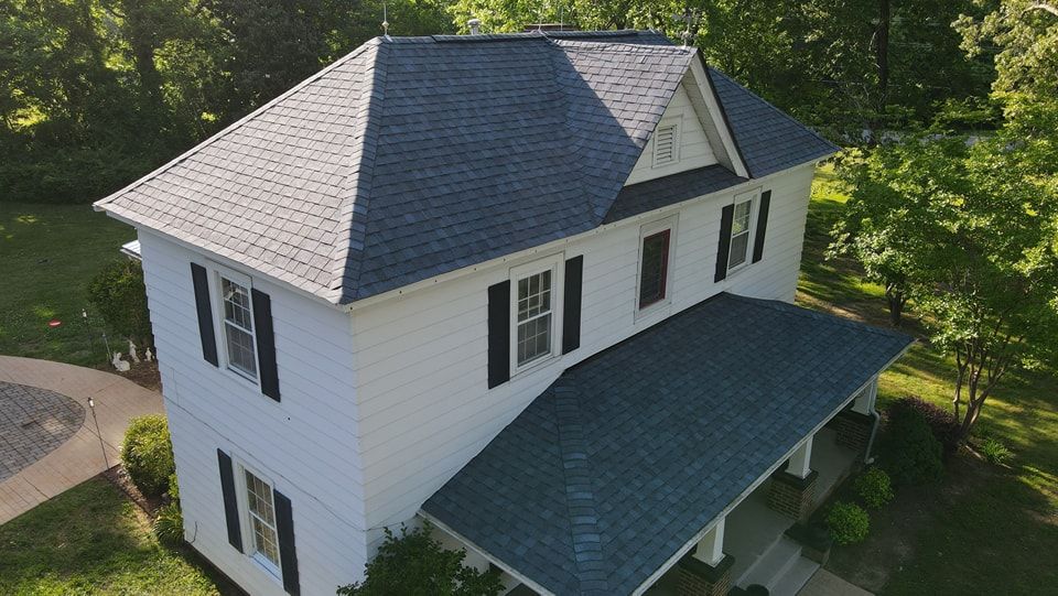 White two-story house with a dark blue roof, black shutters, and porch. Surrounded by trees.