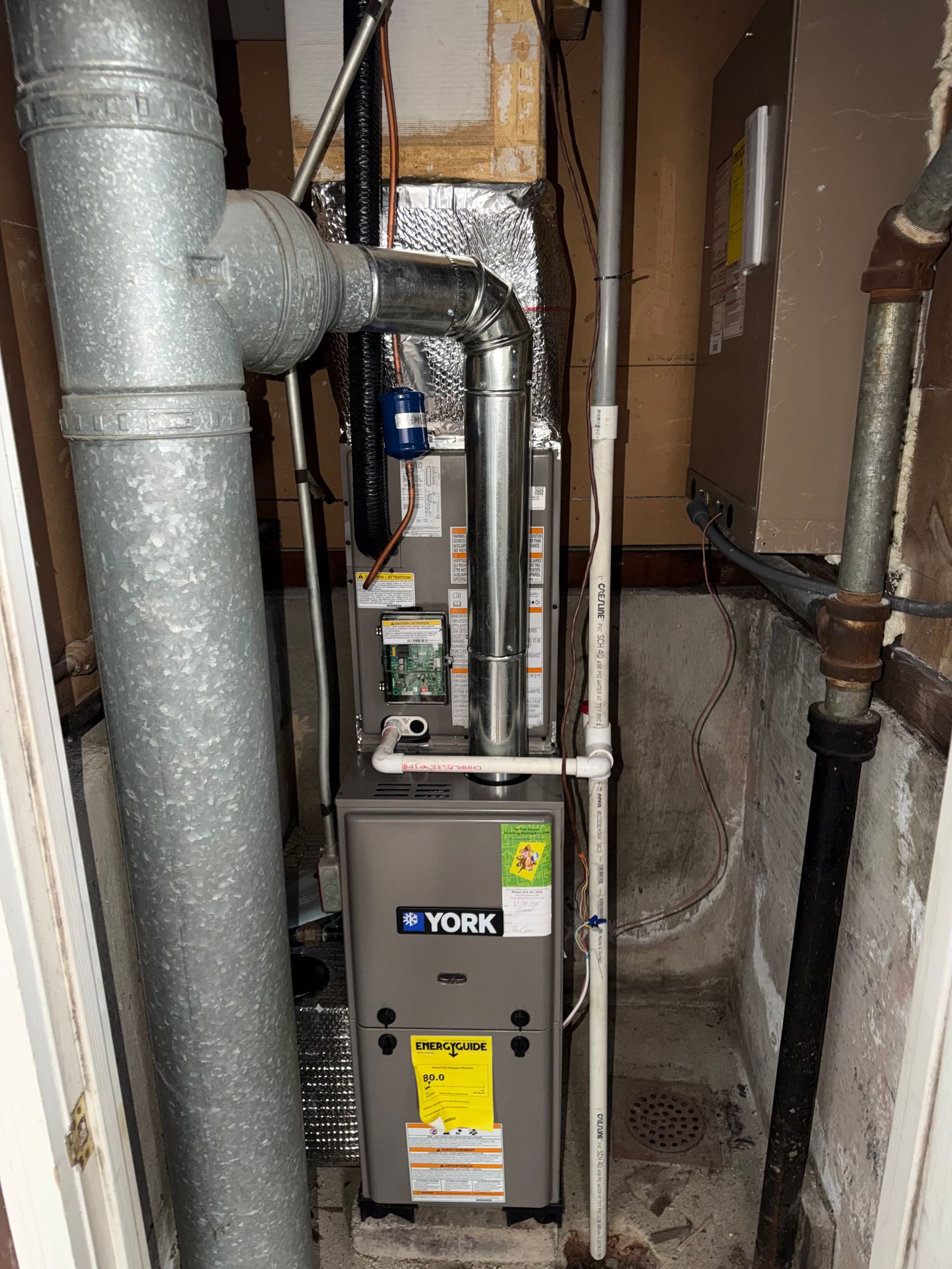 York furnace in a utility closet with exposed pipes and ductwork on either side.