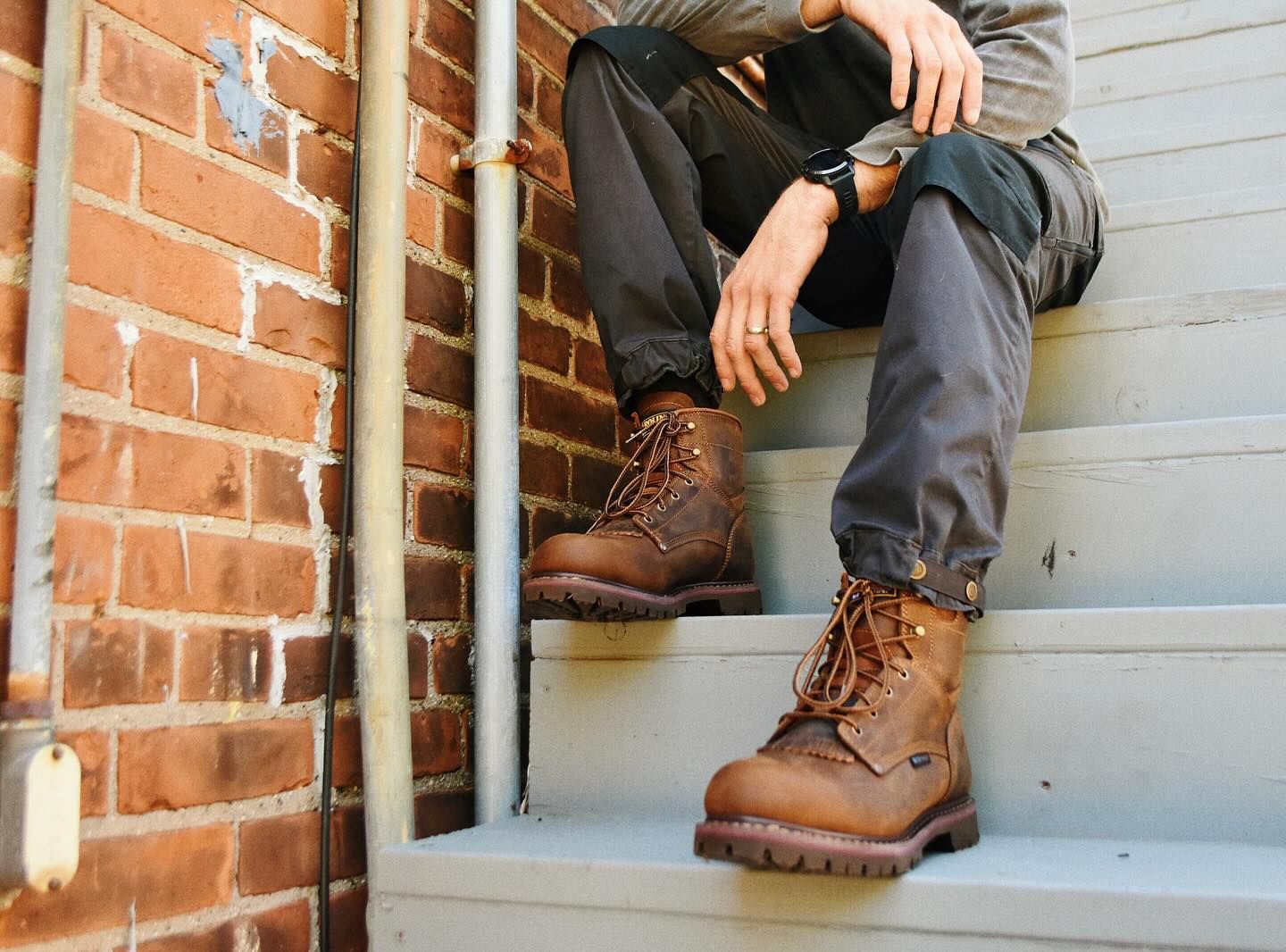 Person in work boots and dark pants sits on outdoor stairs next to a brick wall.