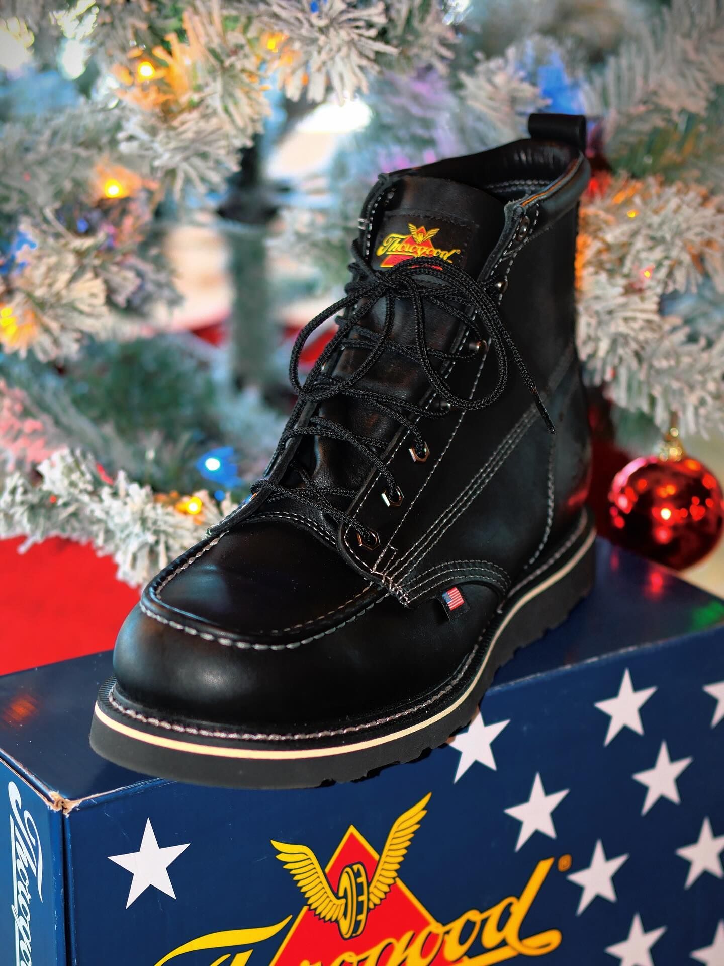 Black work boot on a box with American flag design, in front of a Christmas tree with ornaments.