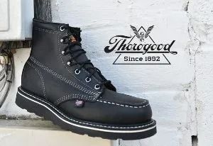 Black Thorogood work boot with white stitching and sole, leaning against a white brick wall.