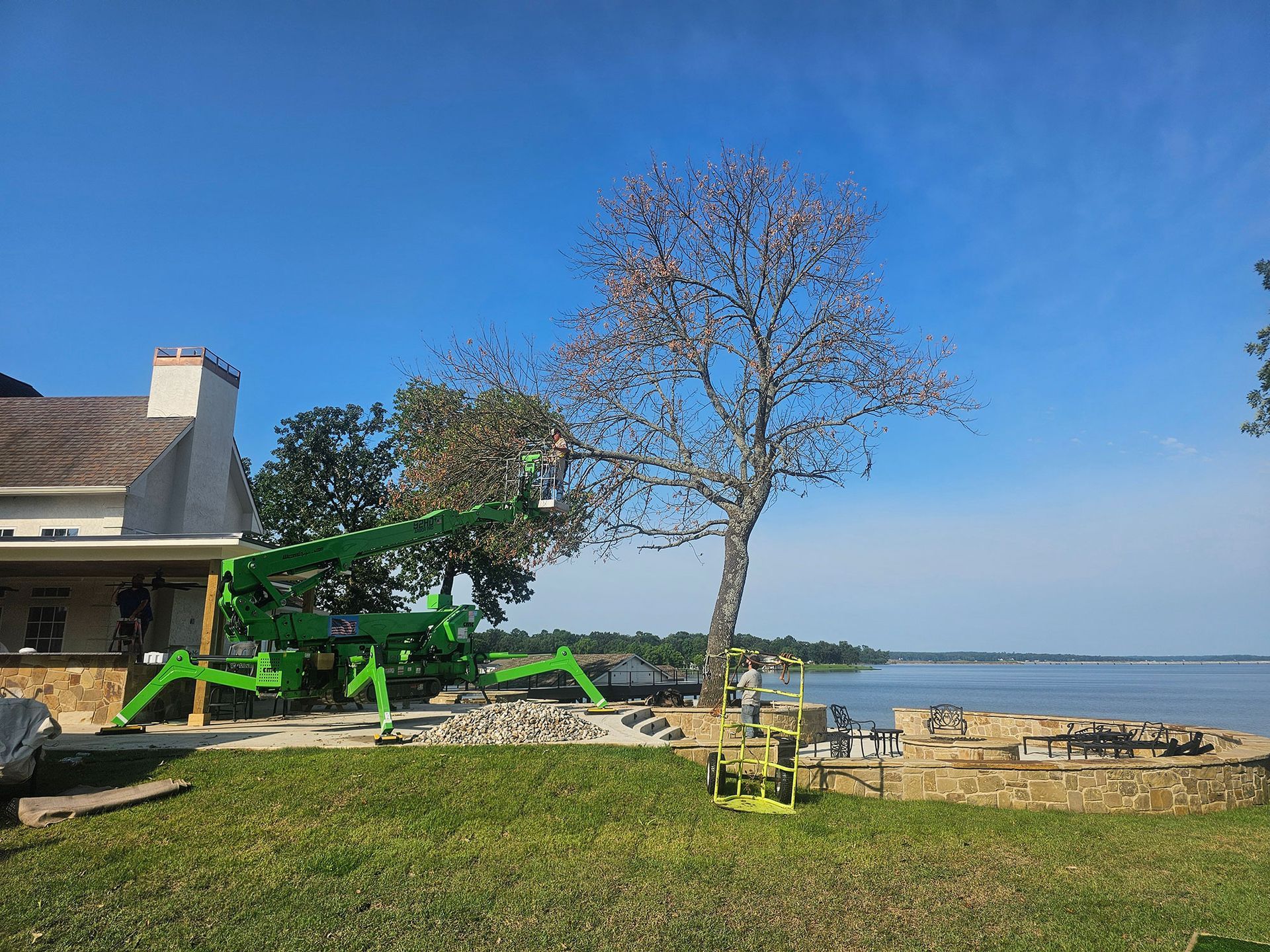 Green crane lifting a tree beside a house in a sunny lakeside yard