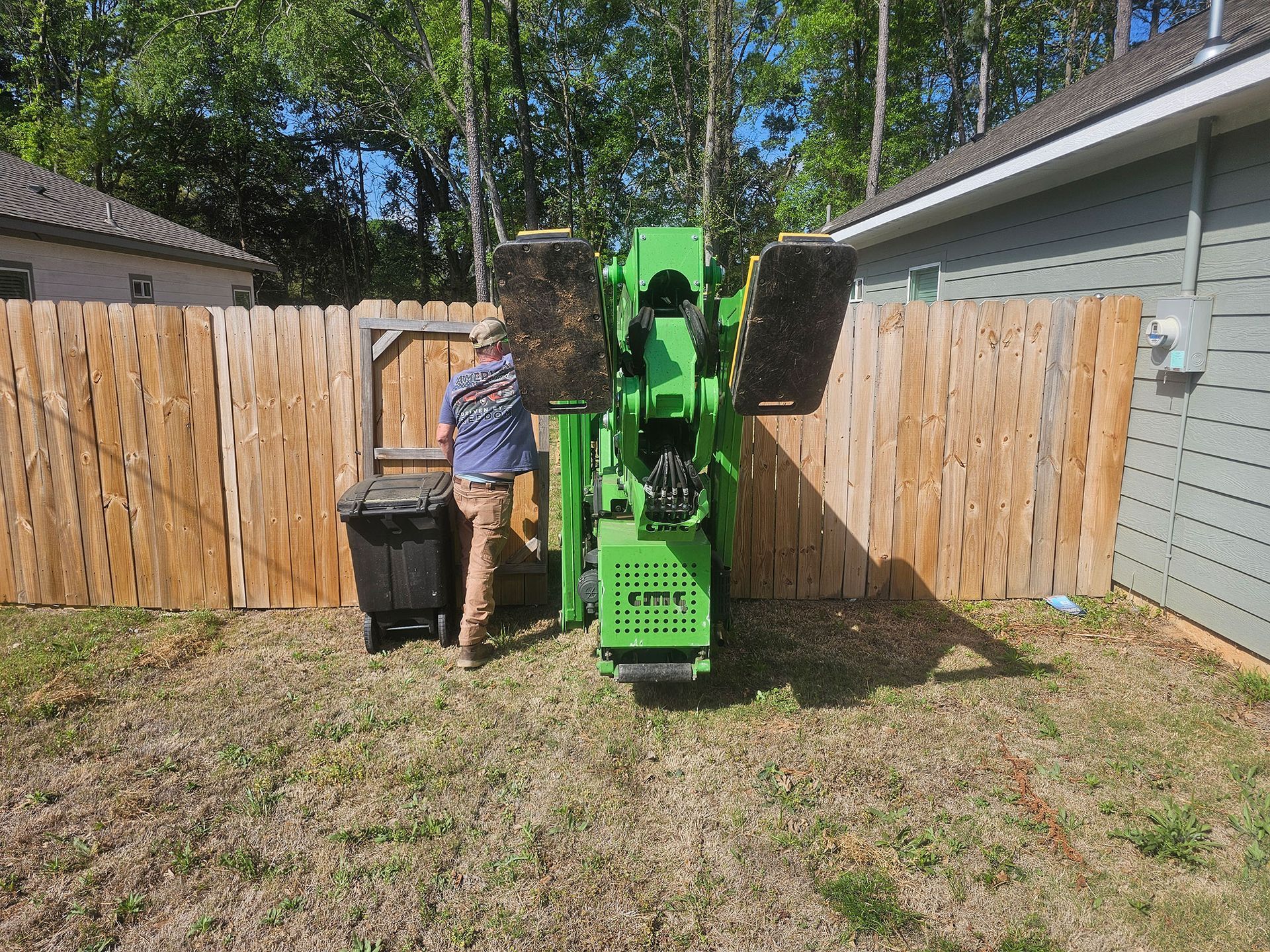 Person beside a green skid steer in a backyard, with a trash bin and wooden fence nearby.