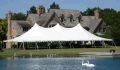 A large white tent is sitting in front of a large house next to a lake.
