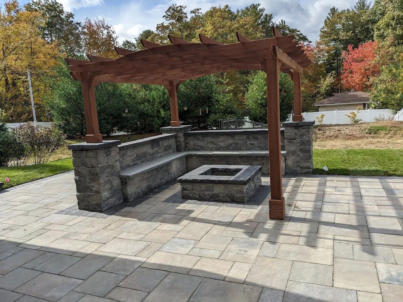 Stone fire pit with built-in bench under a wooden pergola in a paved outdoor garden area