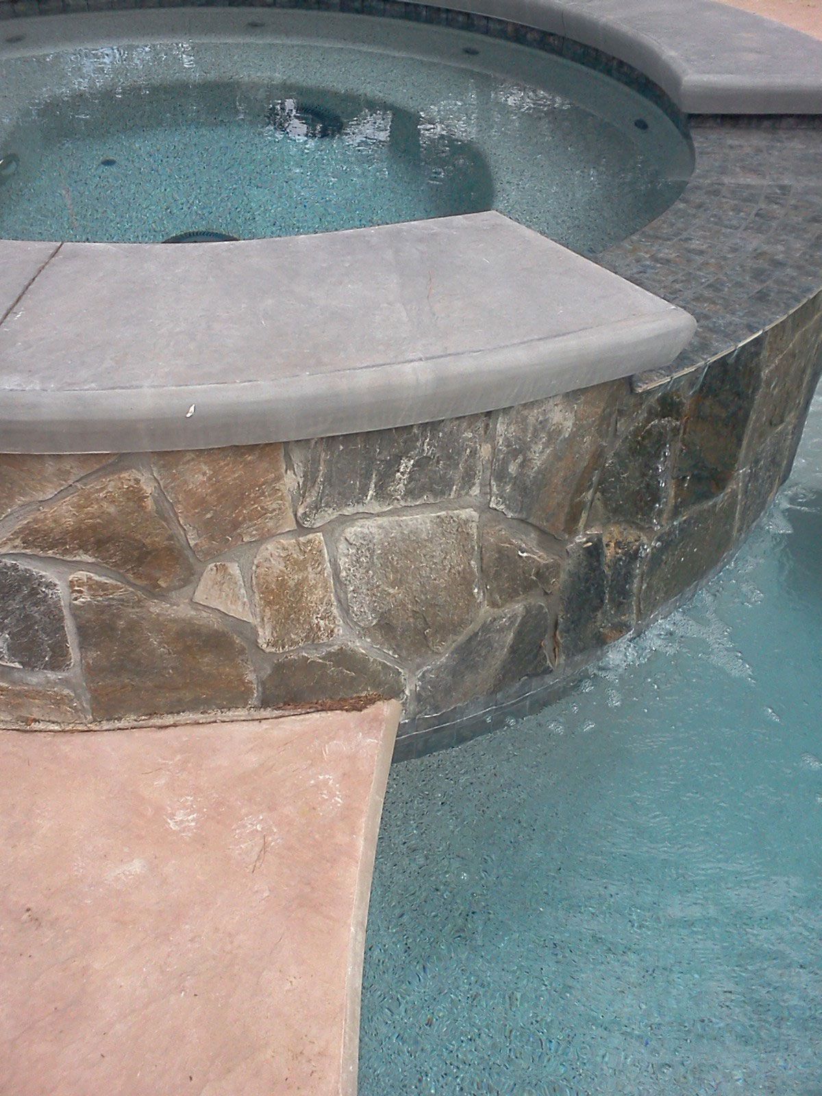 Stone-and-concrete hot tub beside a turquoise swimming pool, with water rippling inside.