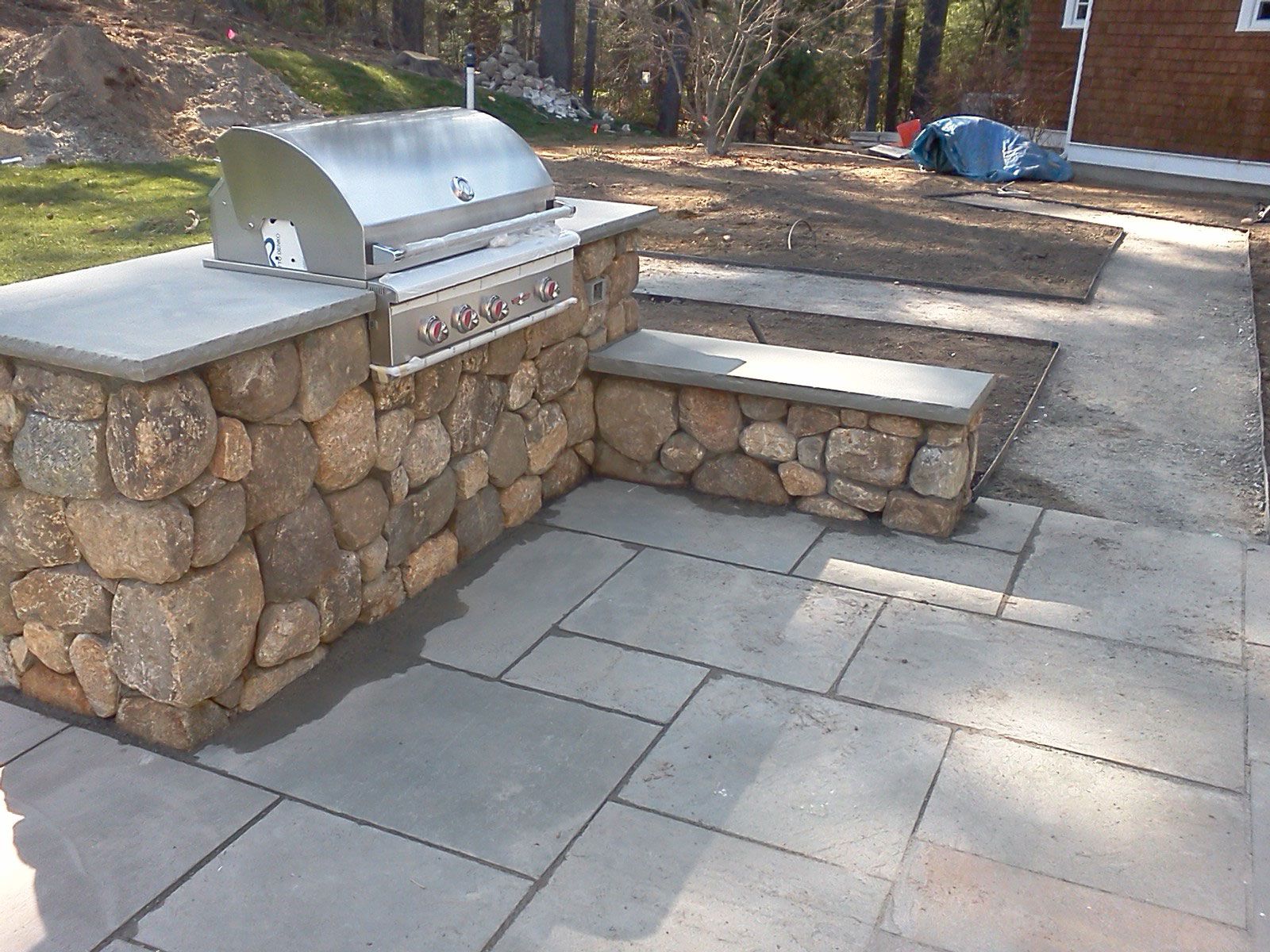 Stone outdoor grill and bench seating area on a paved patio in a park-like setting