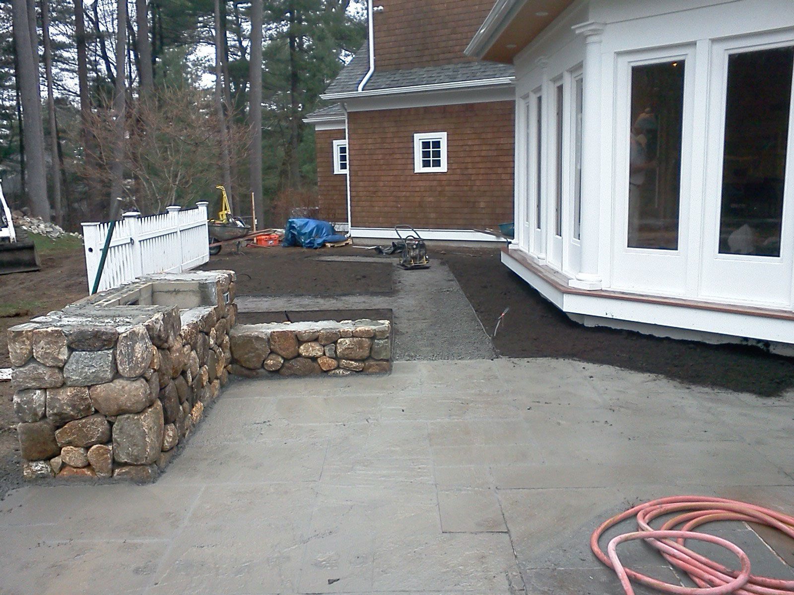 Concrete patio beside white house with stone retaining walls and red hose on the ground