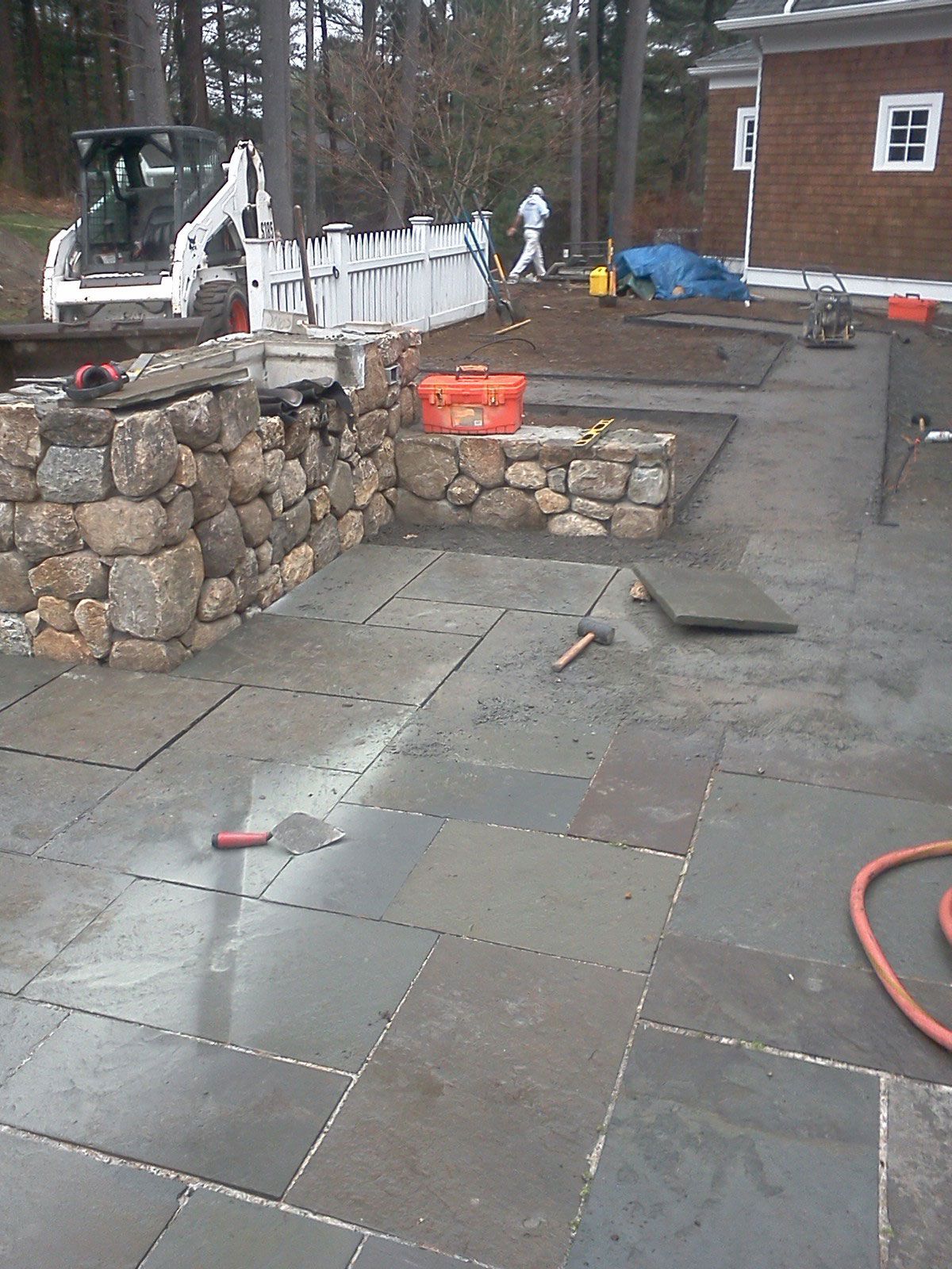 Stone patio under construction with white retaining wall, excavator, and orange hose by a house