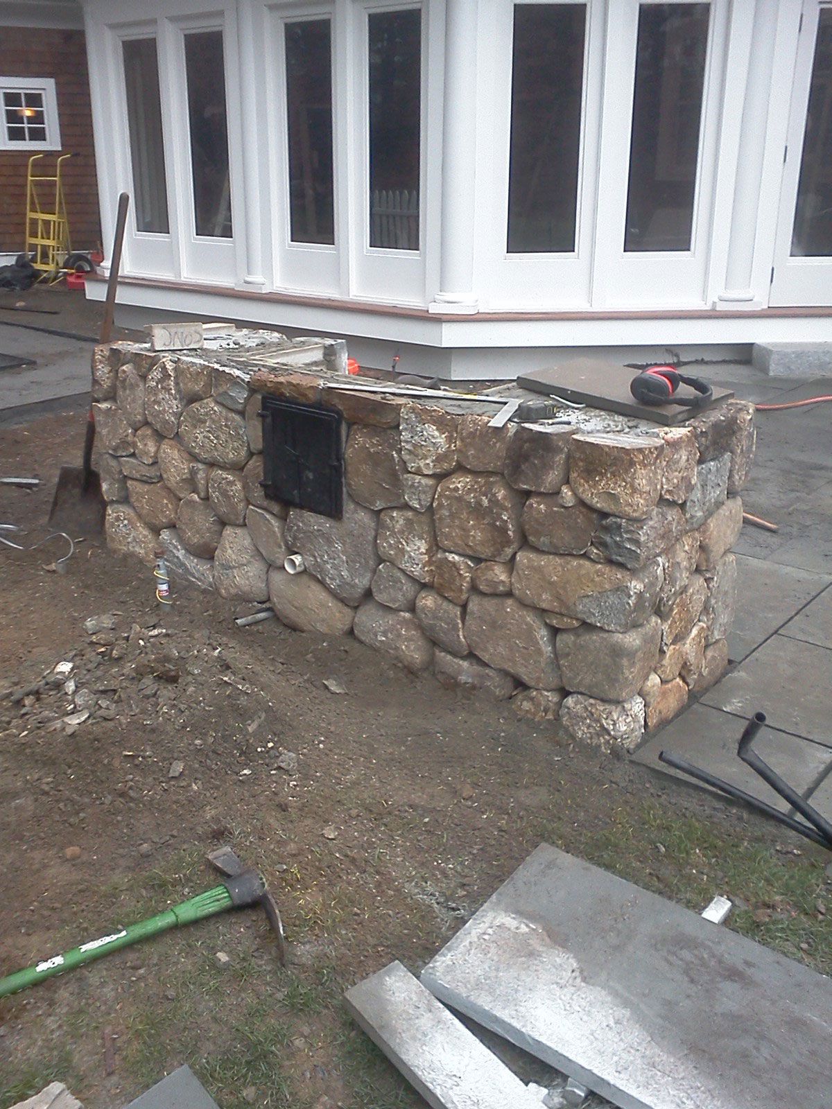 Stone outdoor structure by a white house, with a black metal access door and construction materials nearby