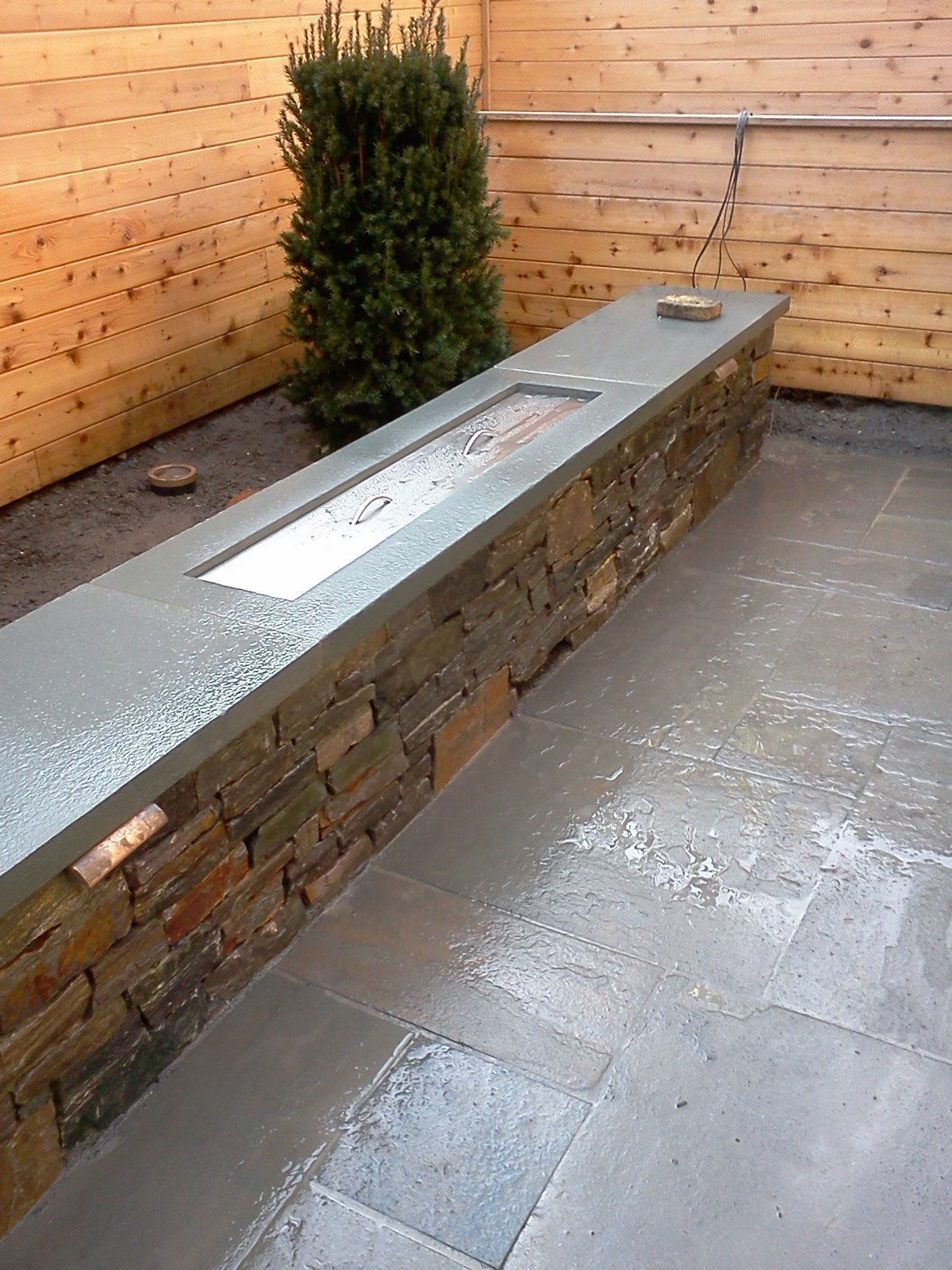 Outdoor stone bench with metal insets beside a wooden wall and small shrub in a paved courtyard
