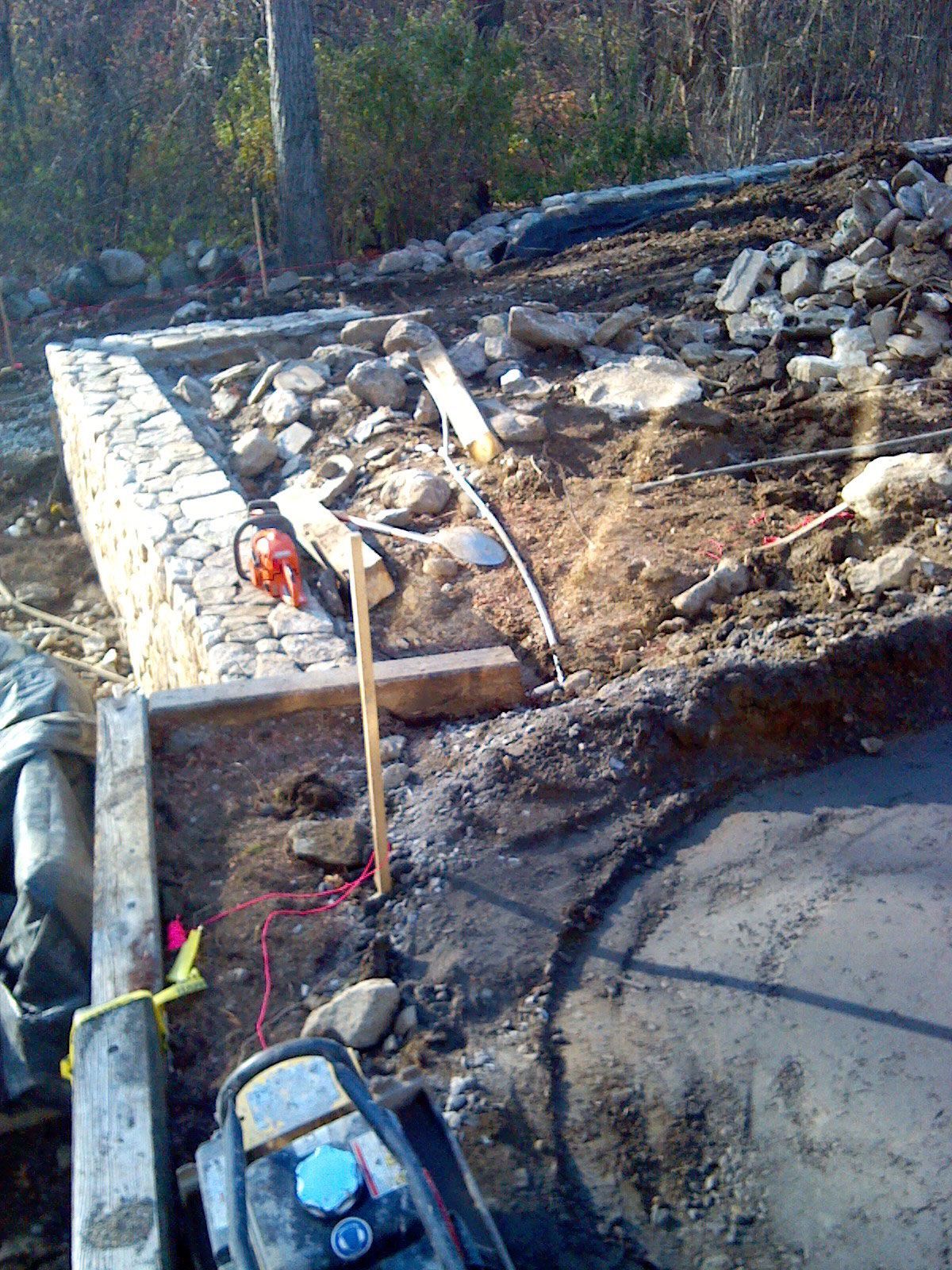 Construction site with gravel, wooden formwork, a shovel, and a wheelbarrow beside a concrete foundation