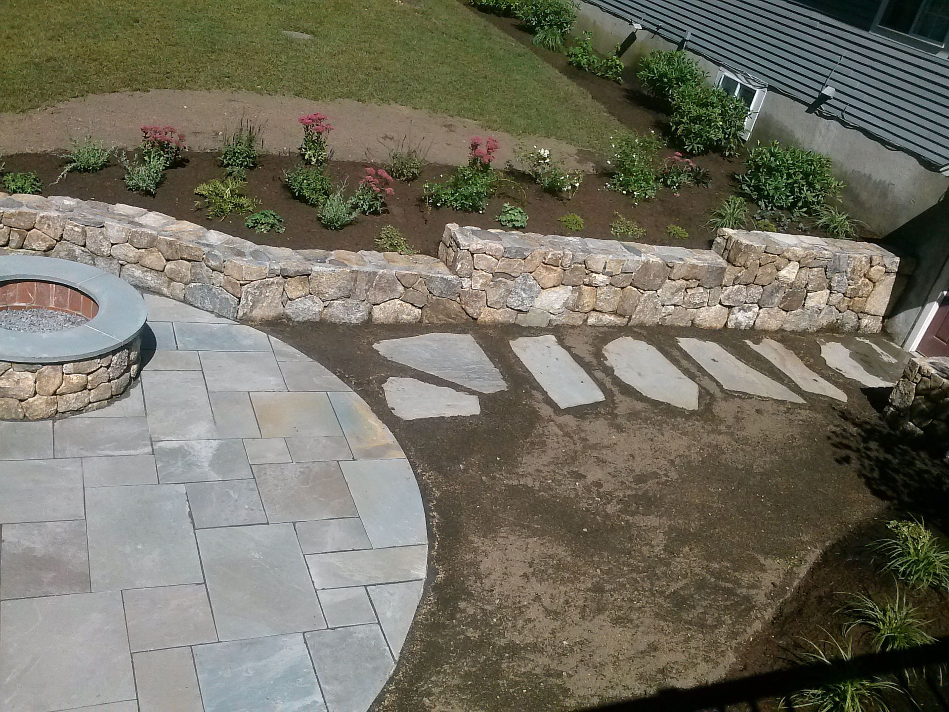 Curved stone patio beside a fire pit, flower bed, and stepping stones in a landscaped yard.