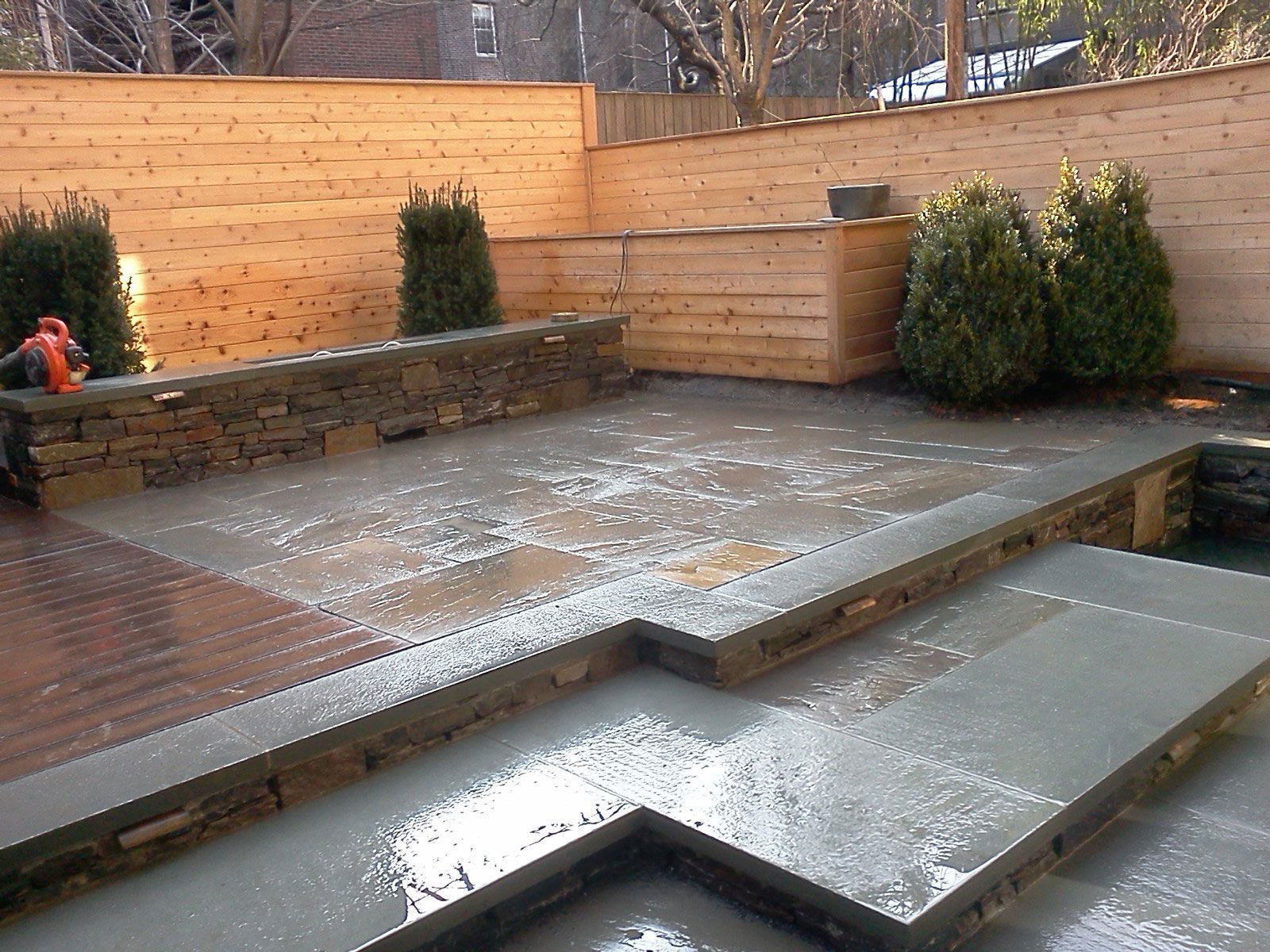 Backyard patio with wet concrete steps, wooden decking, stone wall, and shrubs after rain