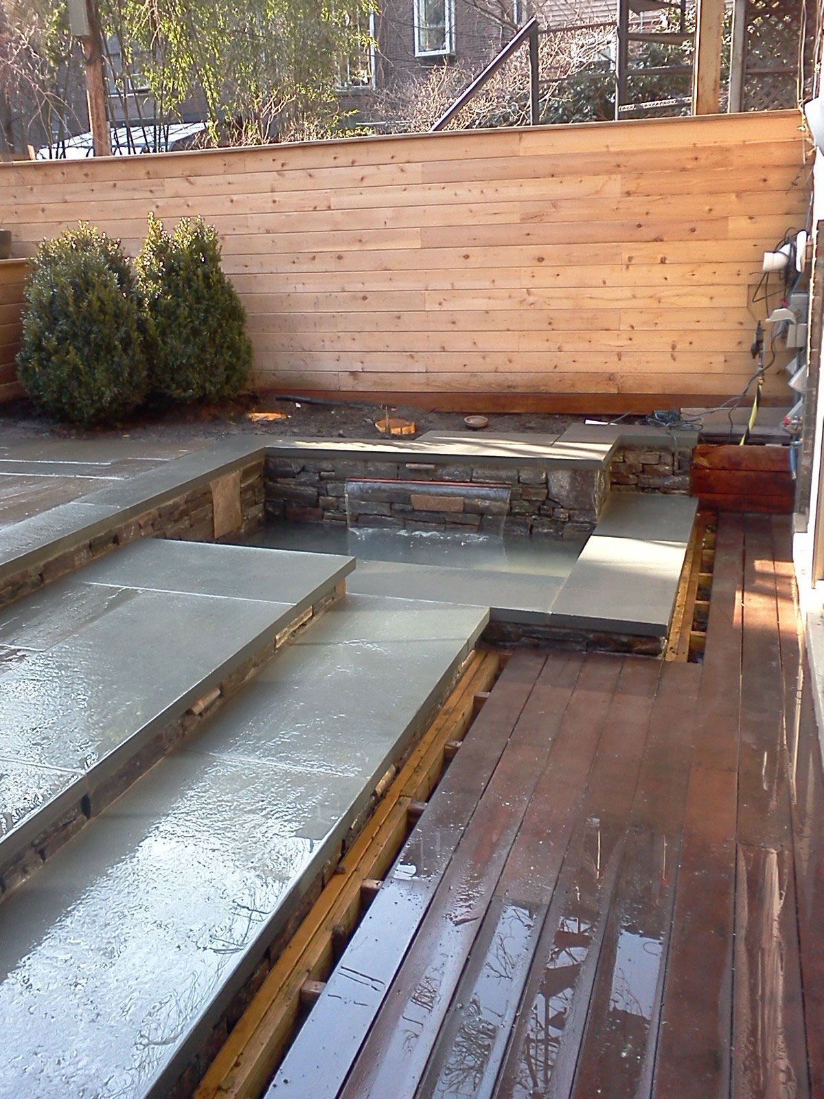 Modern backyard patio with wet wooden deck, concrete steps, stone wall, and shrubs