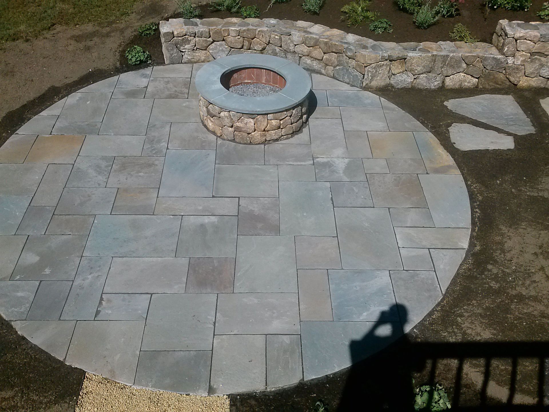 Paved stone patio with a circular fire pit and stone retaining wall in a backyard garden.