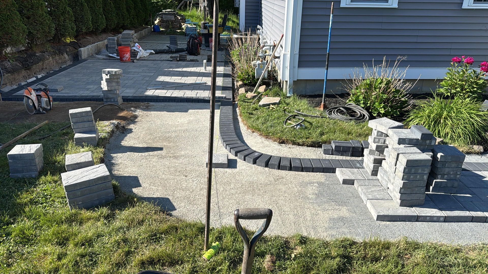 Stone patio and walkway beside a house, with garden beds, stepping stones, and a shovel in the foreground.