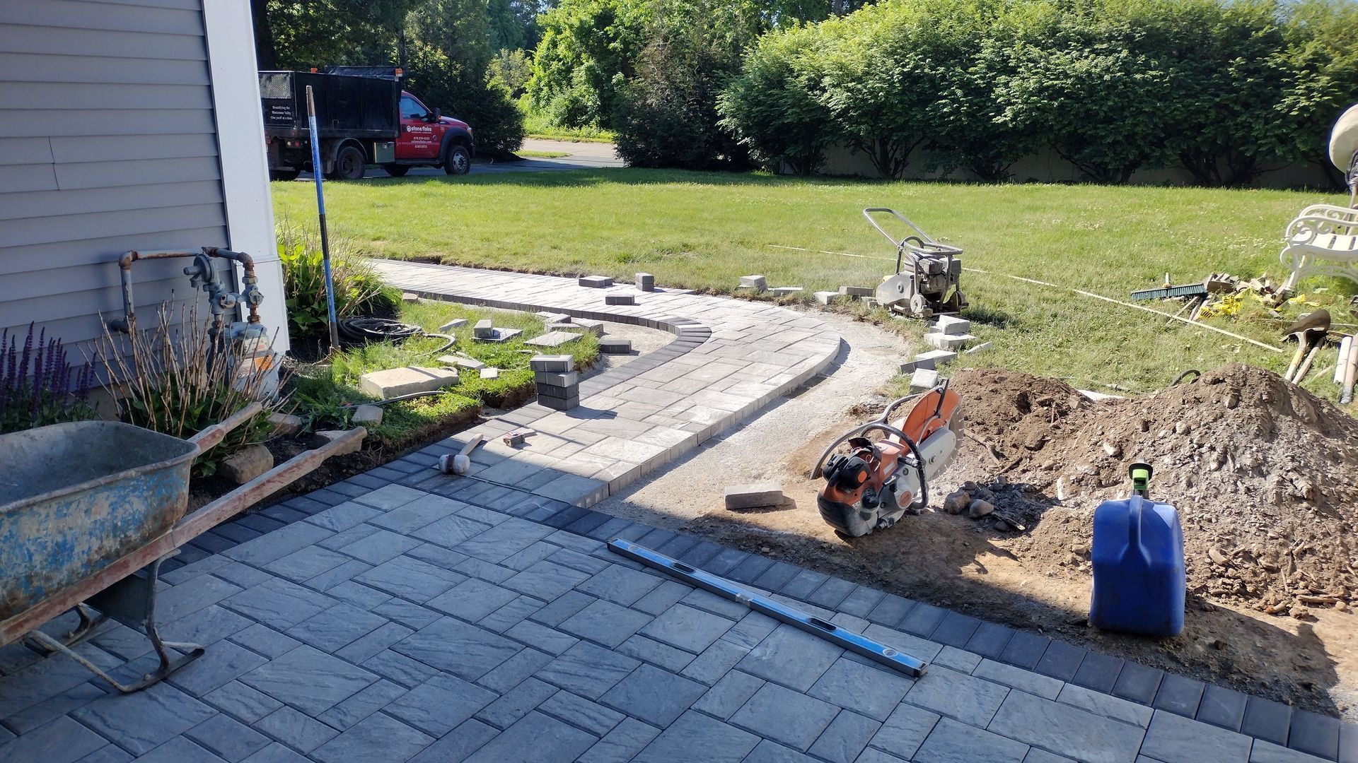 Backyard patio with stone pavers, curved walkway, dirt trench, wheelbarrow, and blue gas can beside a lawn