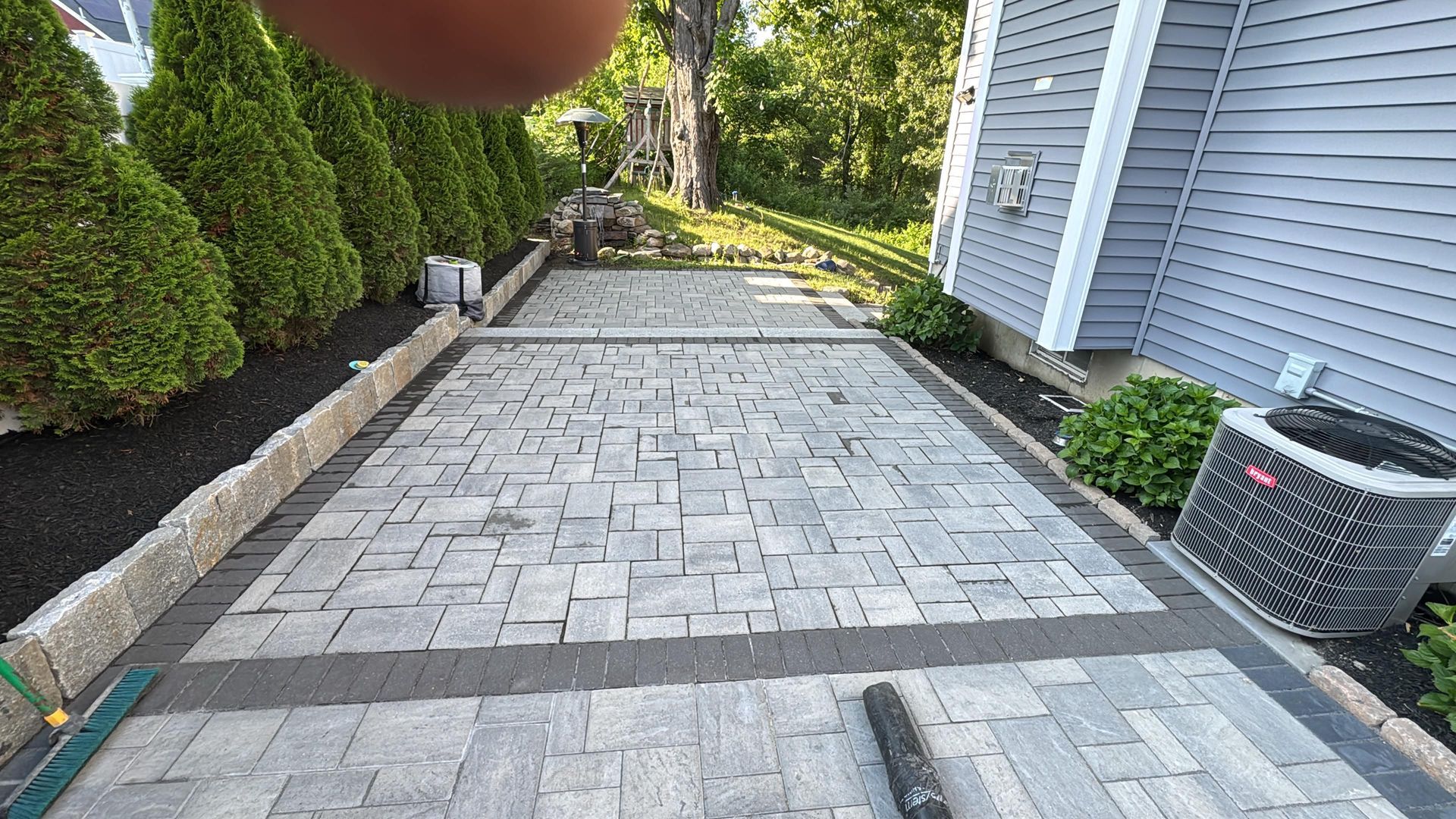 Stone walkway beside a house and trimmed hedges, with a fingertip partly covering the top of the view.