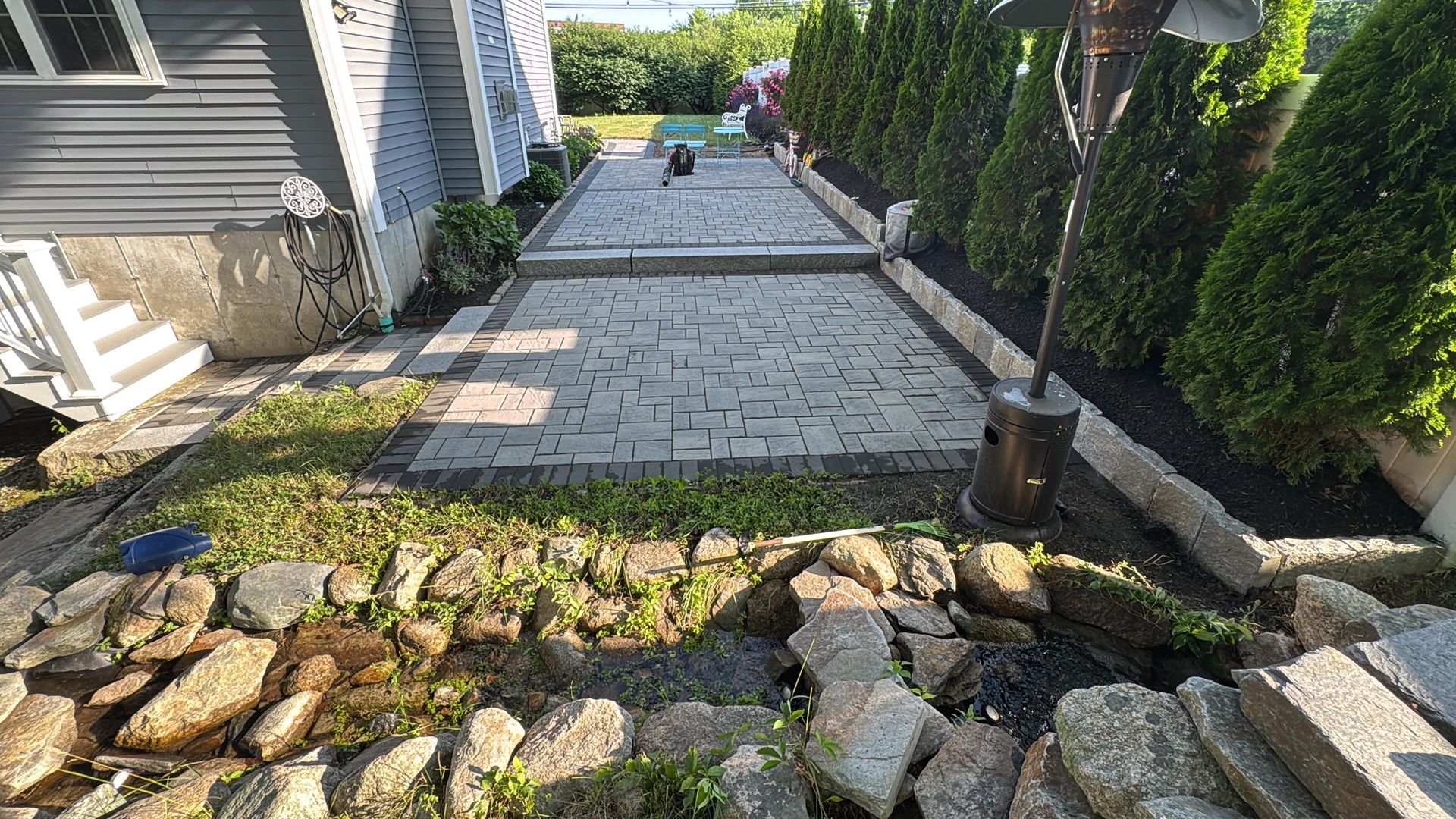 Stone walkway beside a house, bordered by rocks and shrubs, leading to a patio with a small garden light.