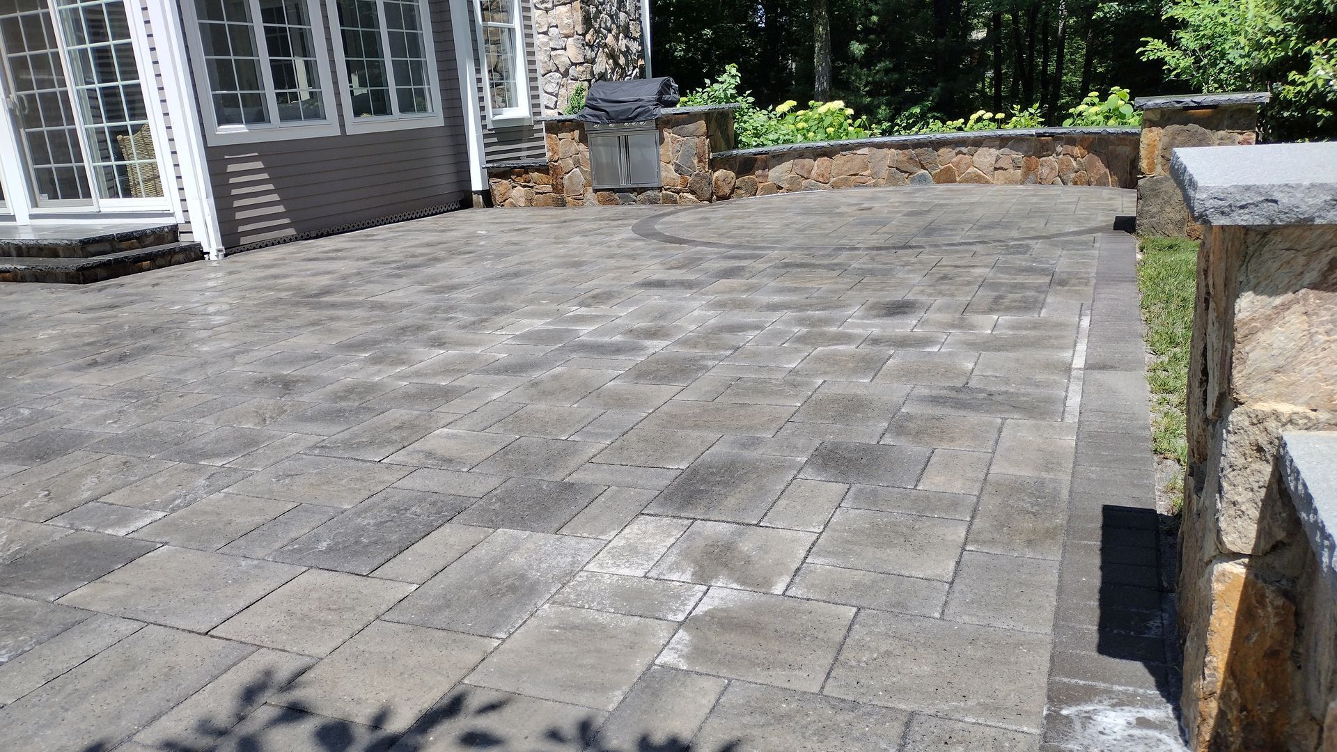 Stone patio with light gray pavers beside a house and stone wall in sunny outdoor setting