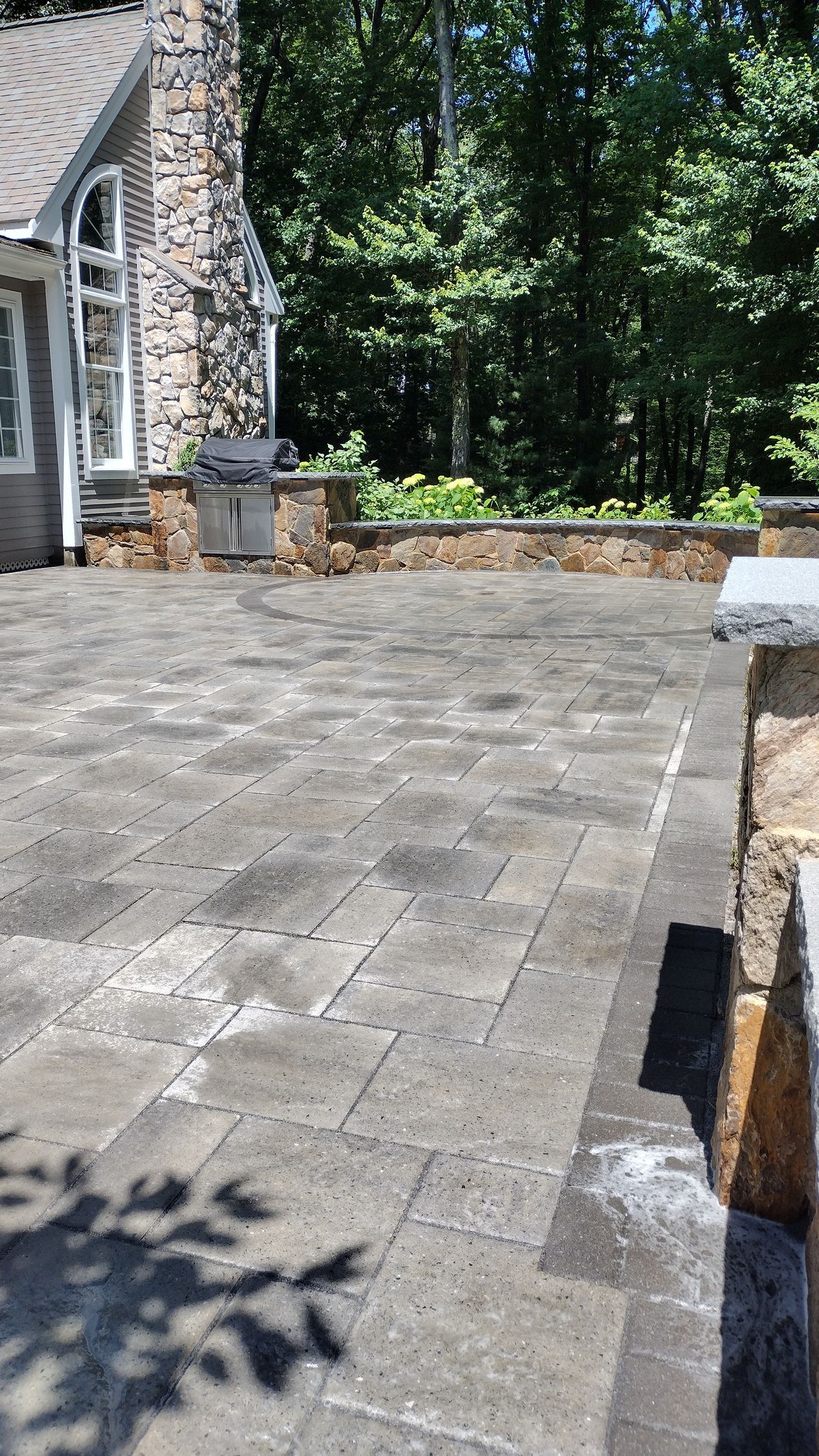 Stone patio beside a house with a grill and trees in the background