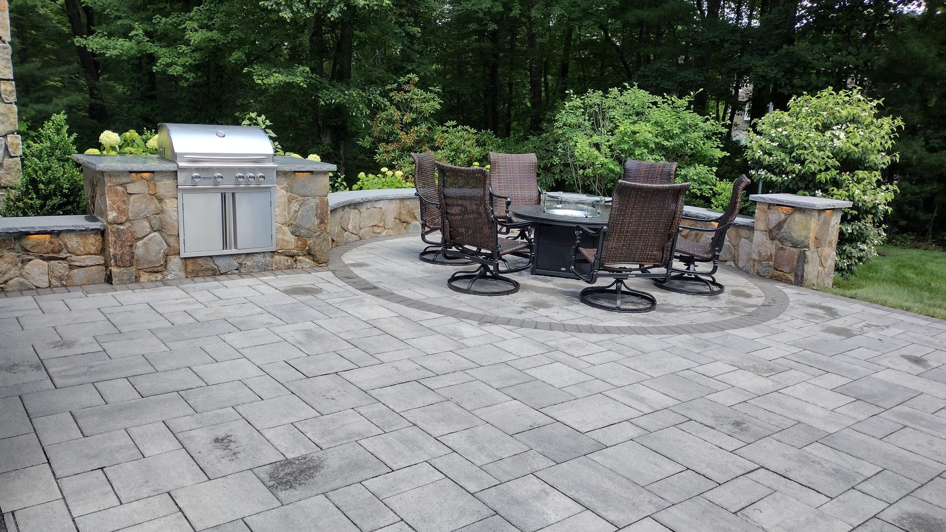 Backyard patio with stone pavers, a grill, and a round table with chairs beside trees and shrubs