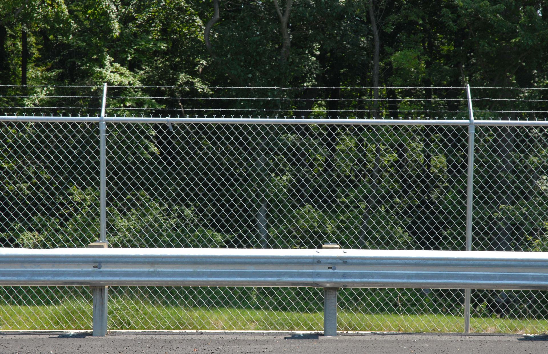 Chain-link fence with barbed wire on top, behind a guardrail, with green trees in the background.