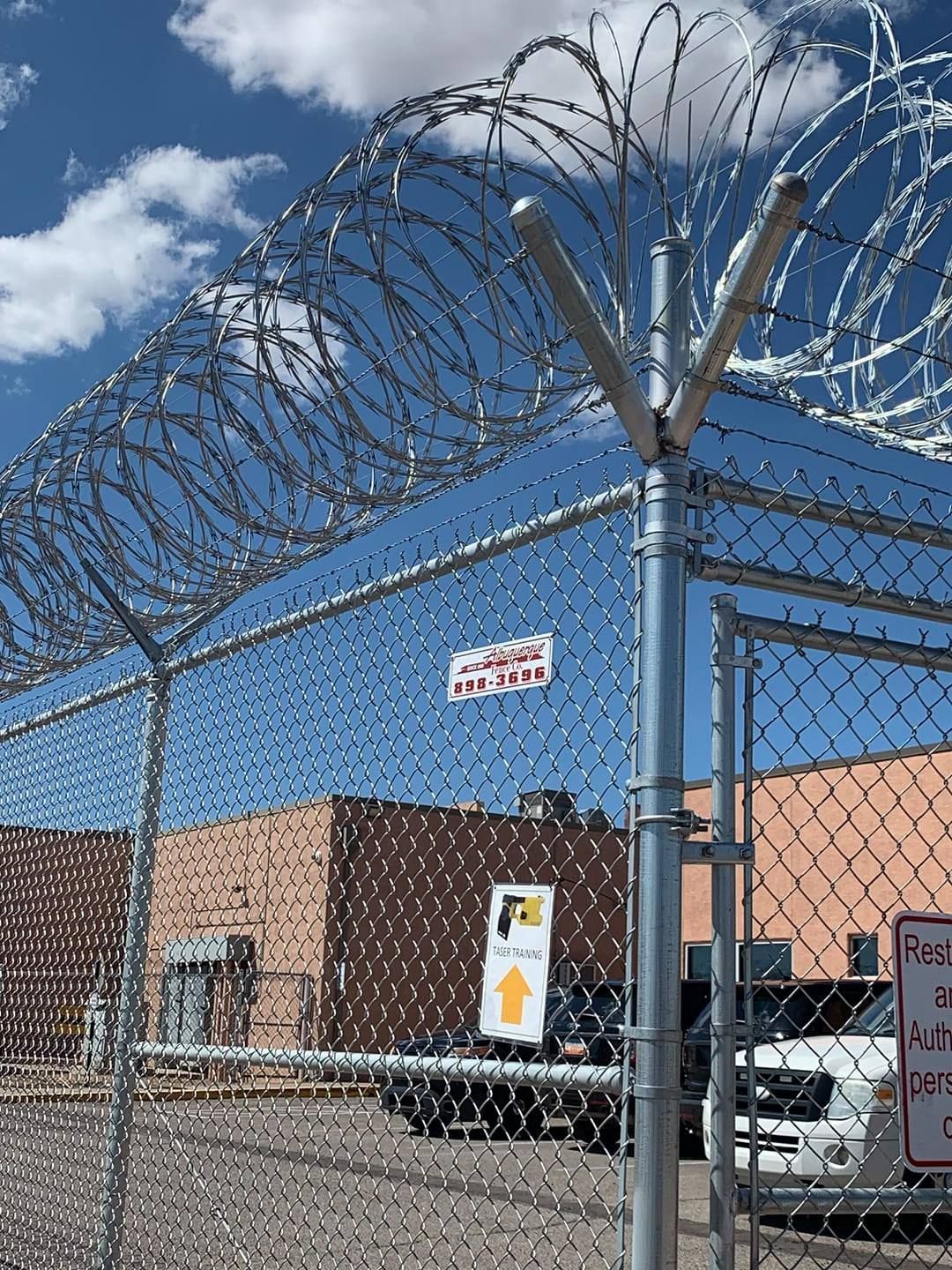 Chain-link fence topped with razor wire. Building and gate with sign in background under a cloudy blue sky.