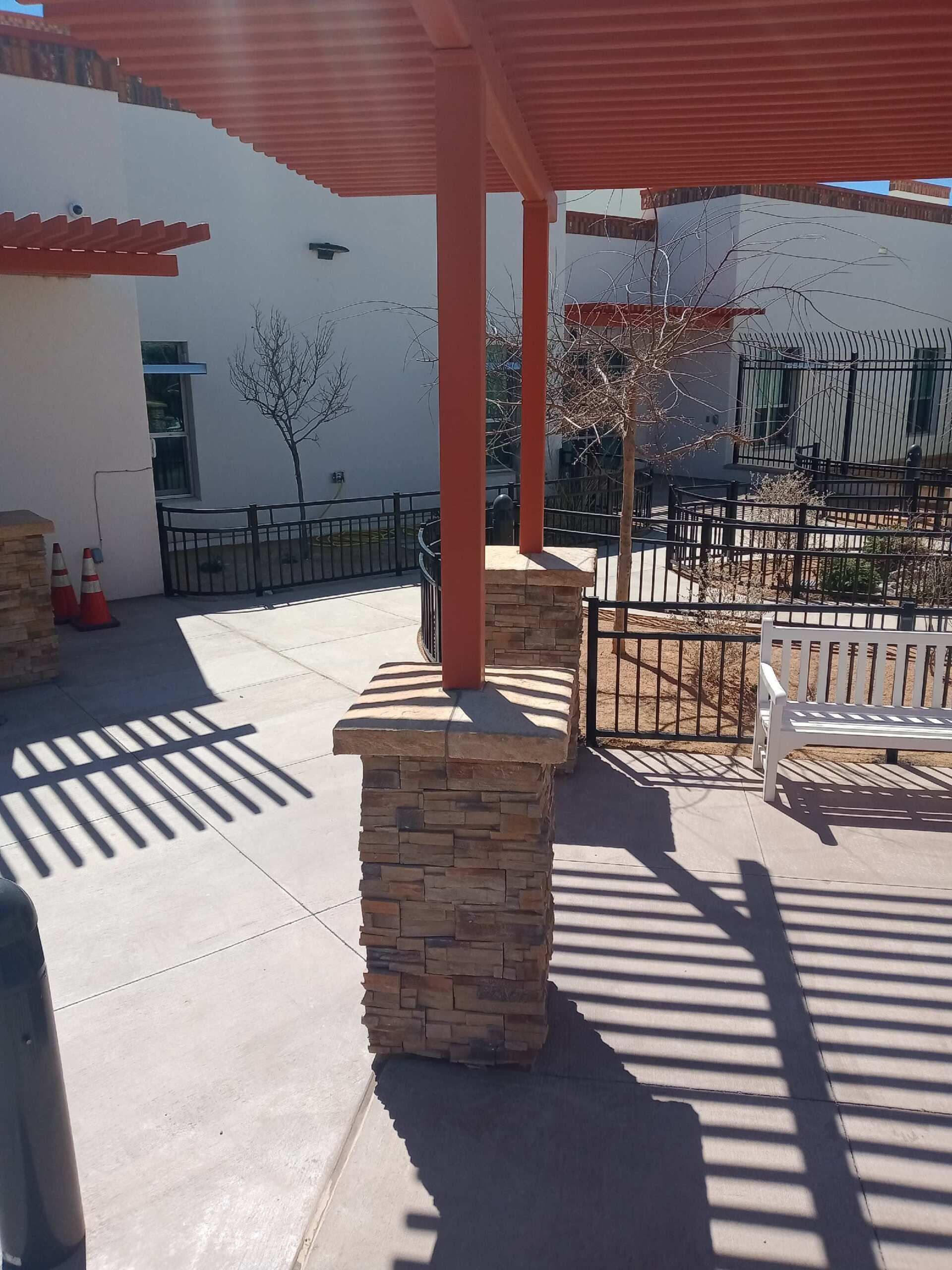 Outdoor patio with red pergola and stone columns, shadows on concrete, and a white bench.