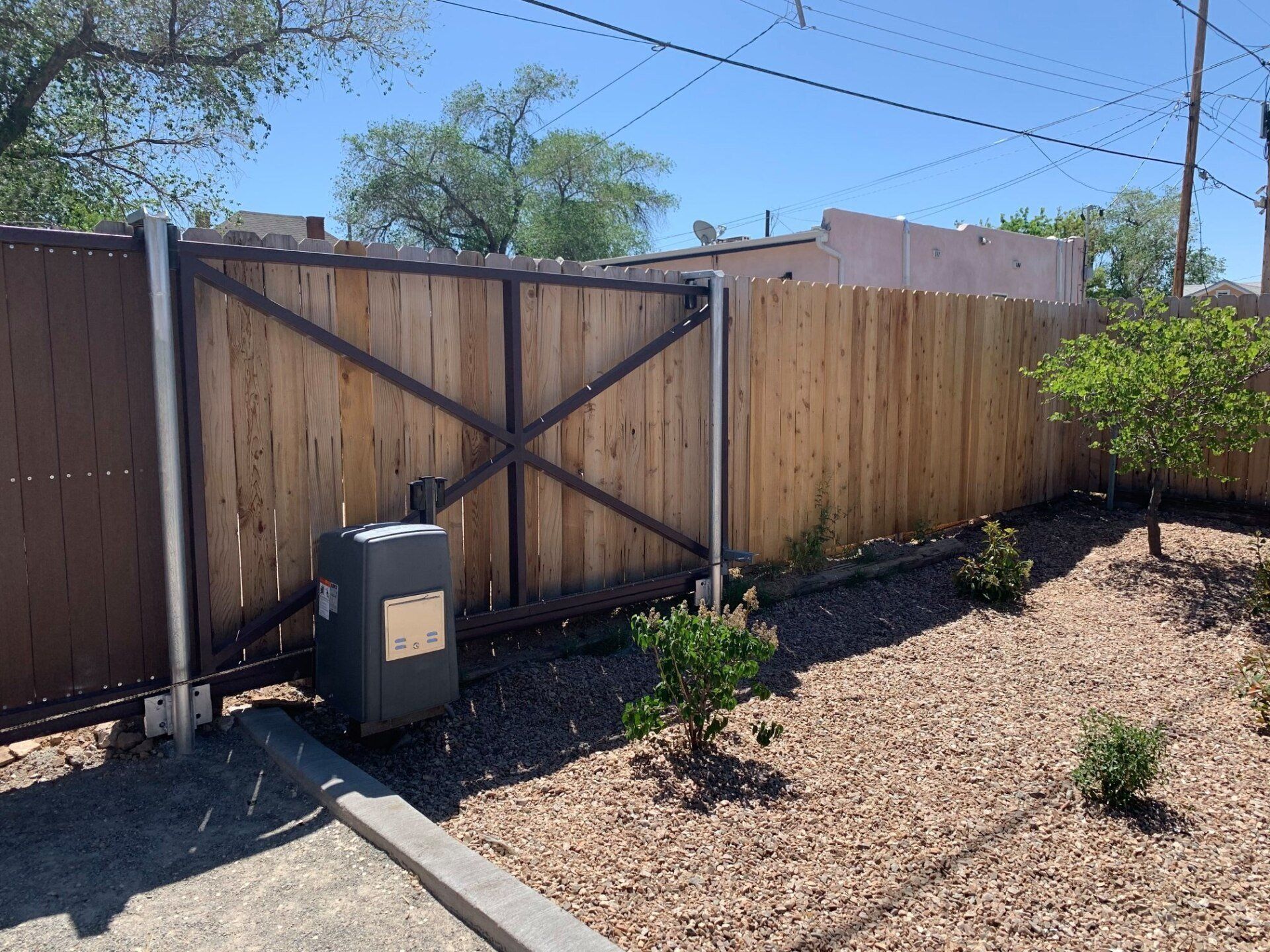 Wooden gate and fence in a backyard; gravel ground with a small bush and gate opener.