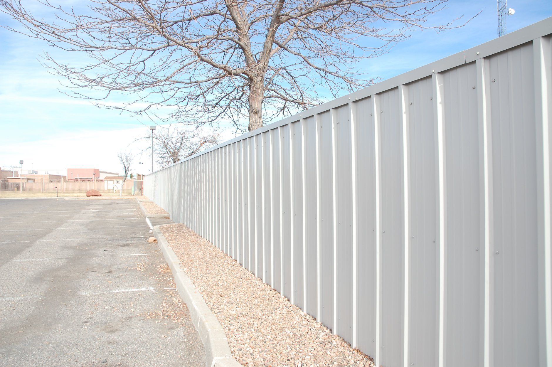 Gray corrugated metal fence along a gravel area beside a paved road with a bare tree in the background.