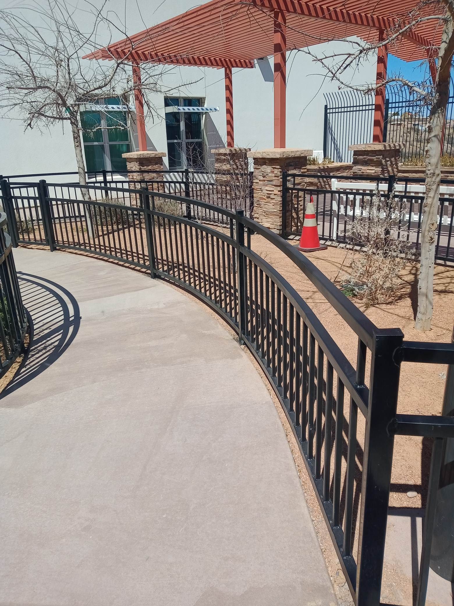 A curved concrete pathway with a black metal fence, leading towards a shaded structure with a red roof.