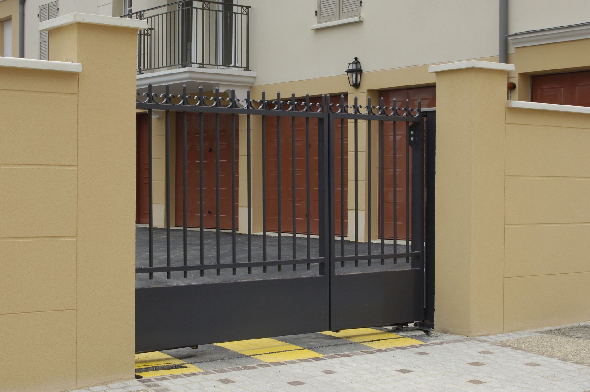 Black metal gate at the entrance to a building. Tan pillars, brown doors in the background. Pavement in front.