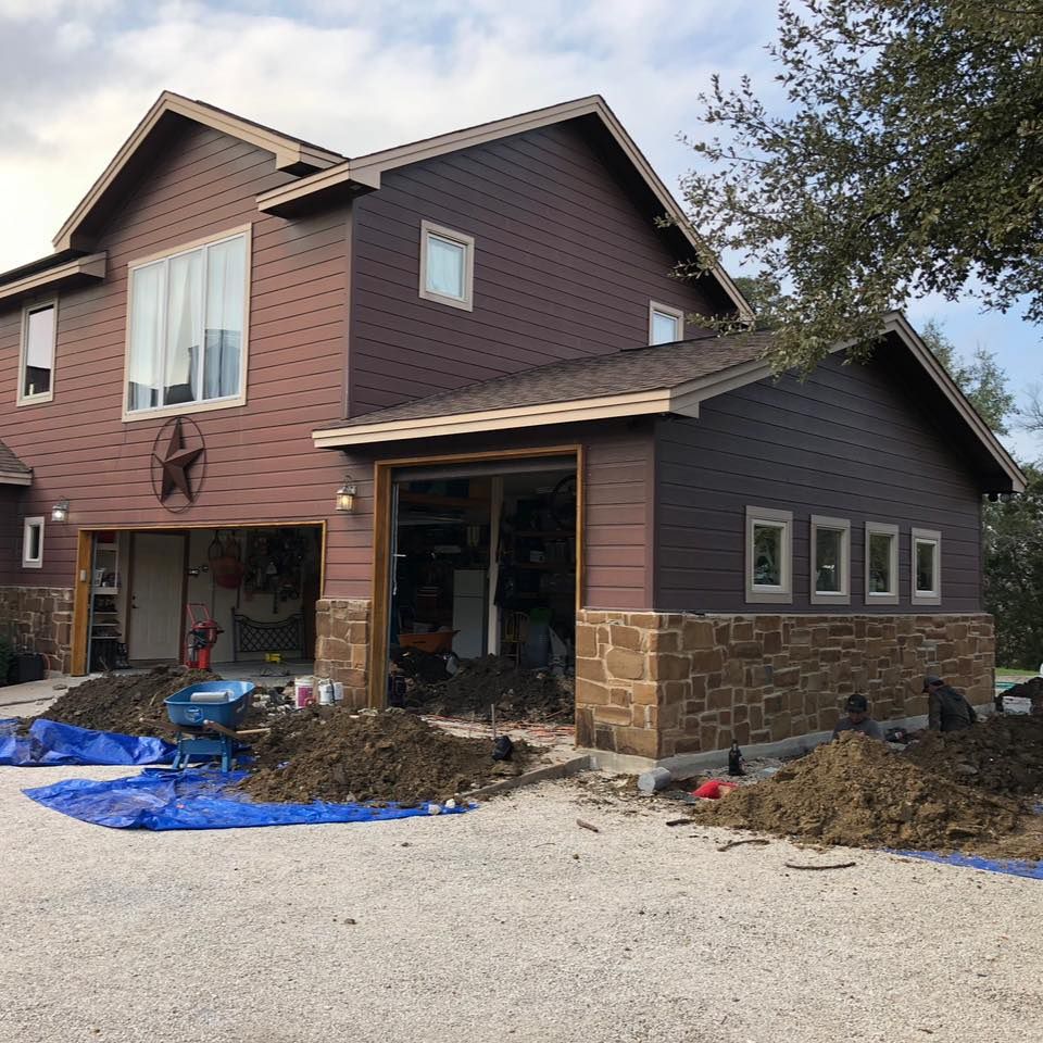Two-story brown house with attached garage under construction, soil piles, blue tarp, and stone facade.