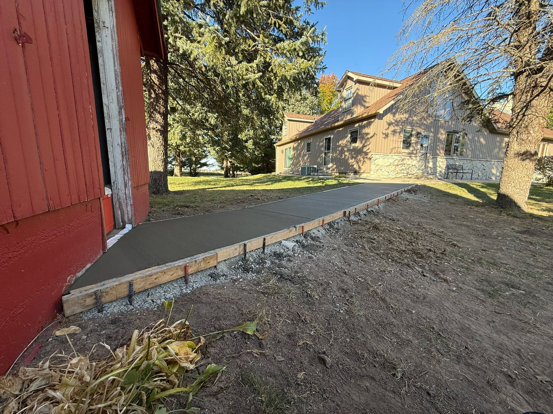 Freshly poured concrete ramp leading up to a red barn. The ramp's formwork is visible, with a stone building and trees in the background.