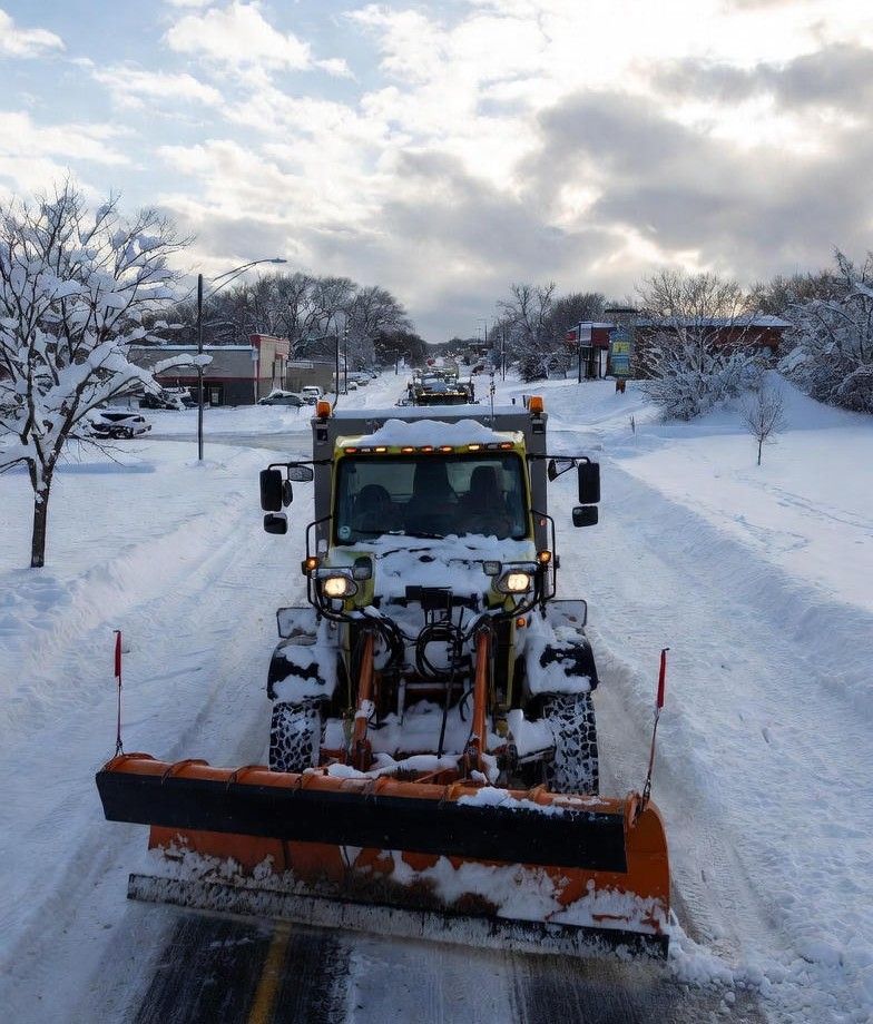 Snowplow clearing a snow-covered street under a cloudy sky. Orange plow blade, yellow lights.
