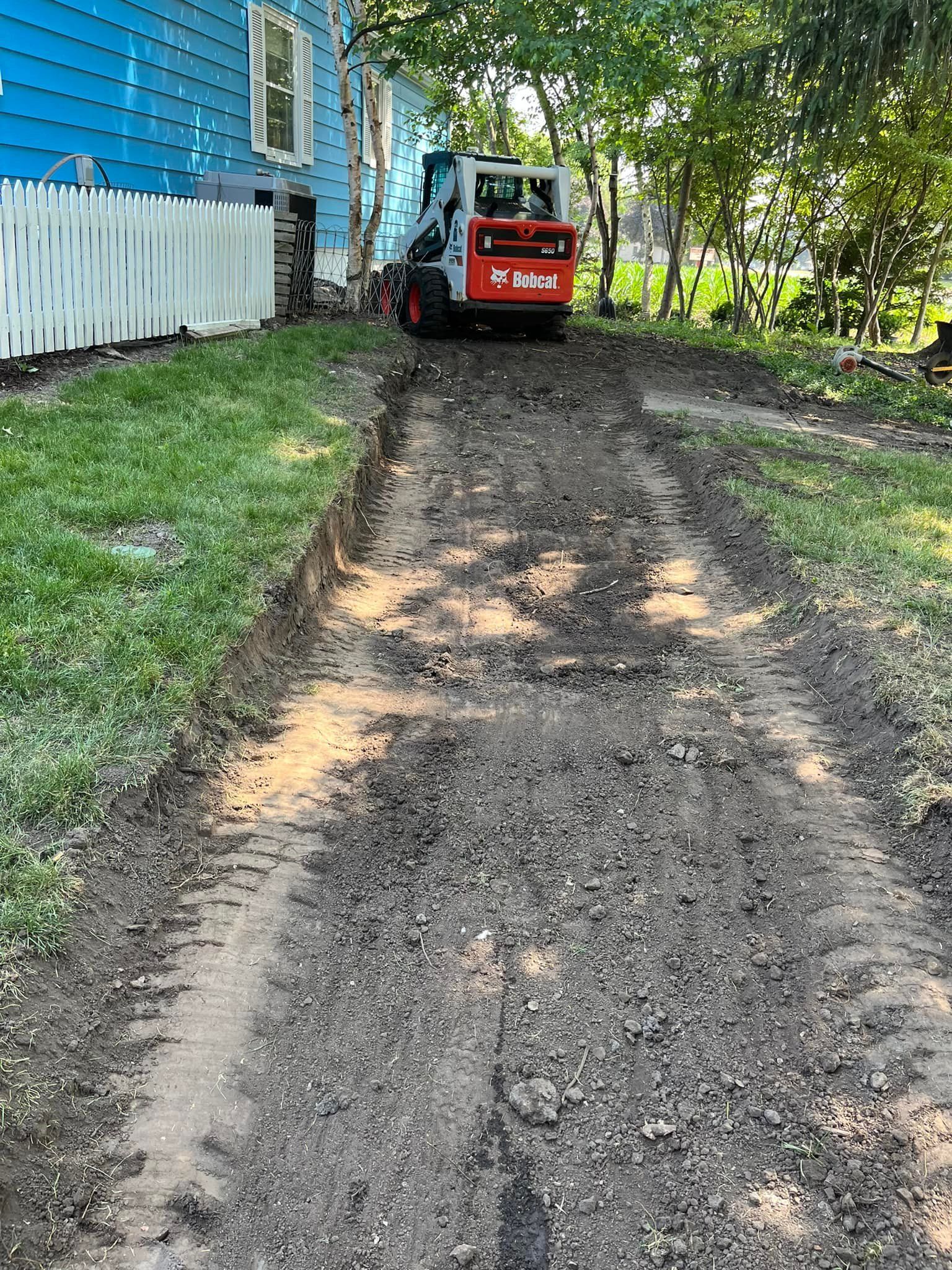 A bobcat is driving down a dirt road next to a house.