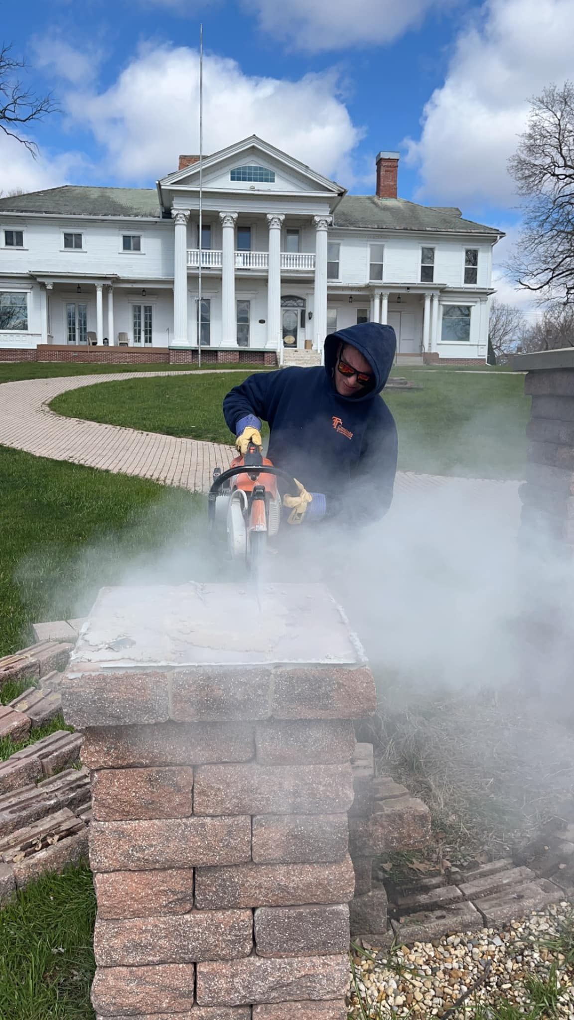 A man is cutting a brick wall with a chainsaw in front of a large house.