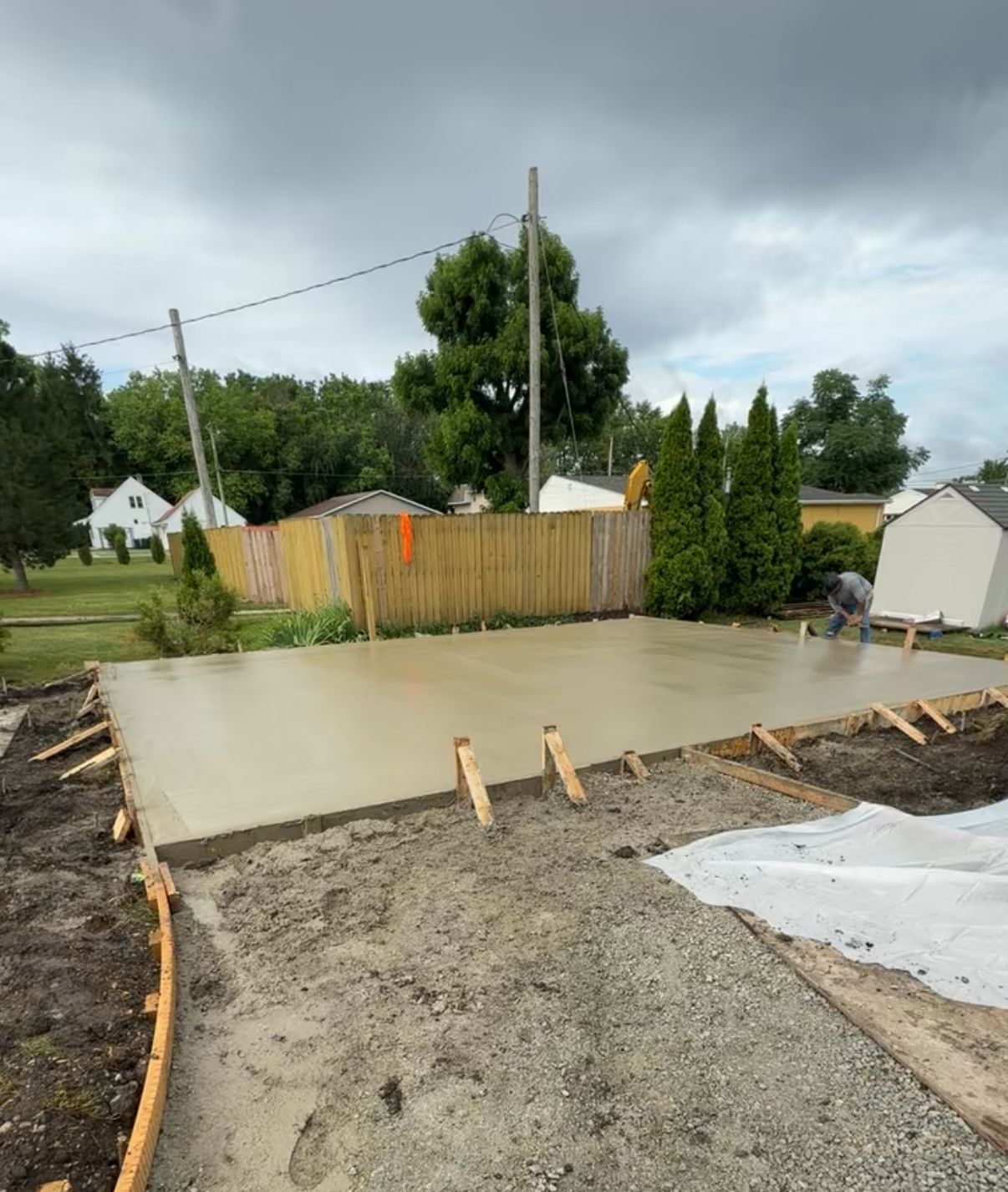 Newly poured concrete slab with wooden formwork, outdoors. Man works on the slab. Cloudy sky.
