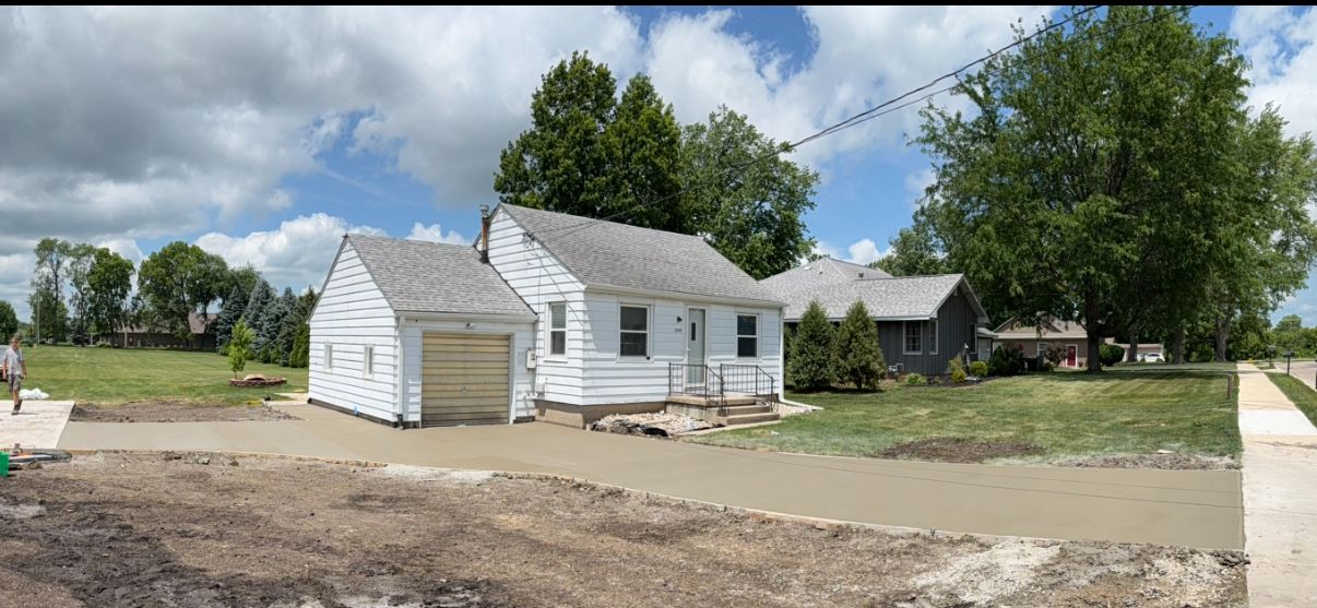 White house with a garage and another house on a cloudy day, dirt and a sidewalk in the foreground.