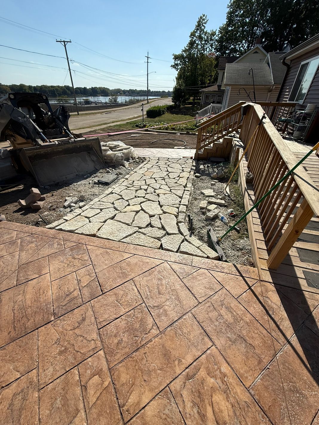 Stone pathway and stamped concrete patio leading to a wooden deck, next to a house with a view of a body of water.