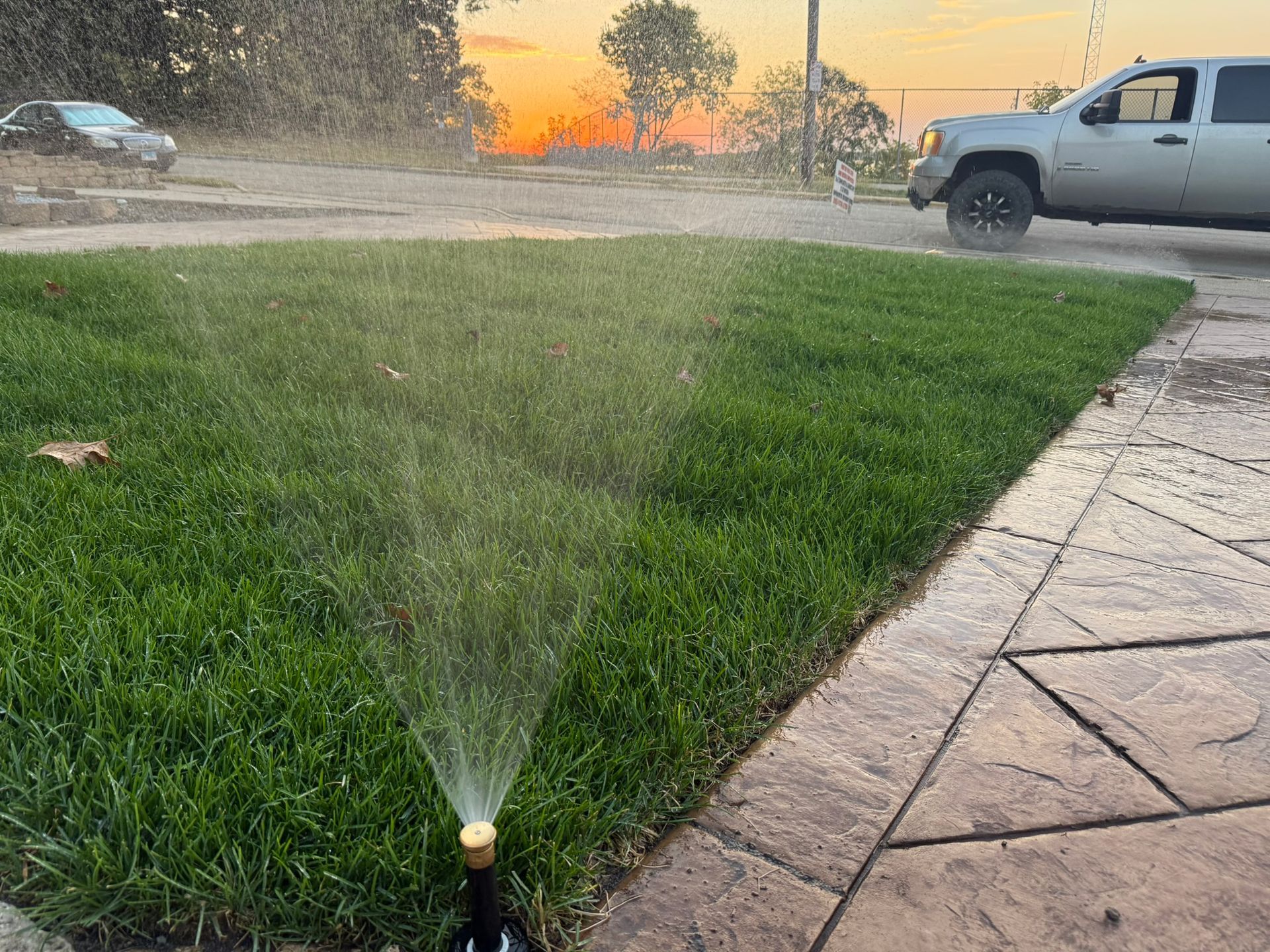 Sprinkler watering a green lawn at dusk, with a truck and car in the background.