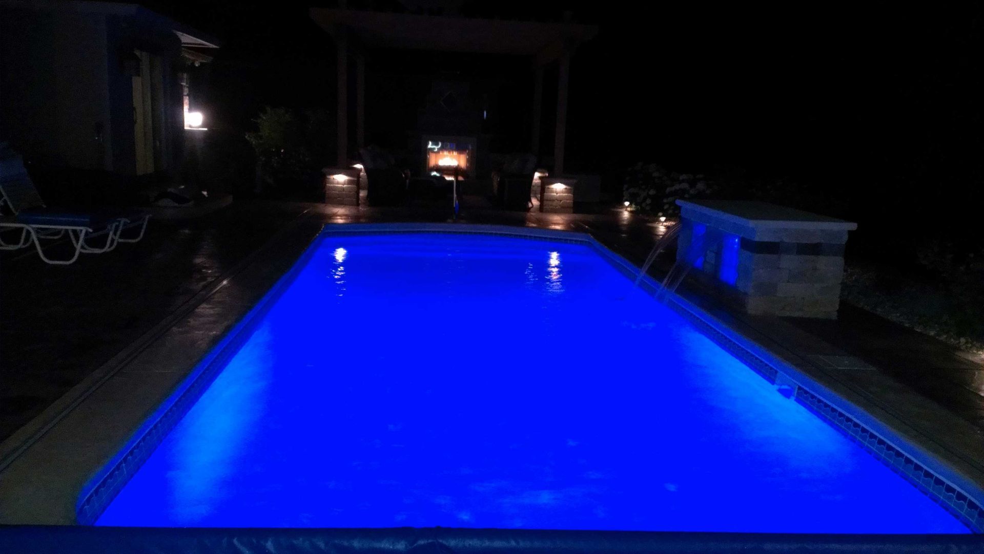 A rectangular swimming pool glows blue at night, surrounded by a dark patio.