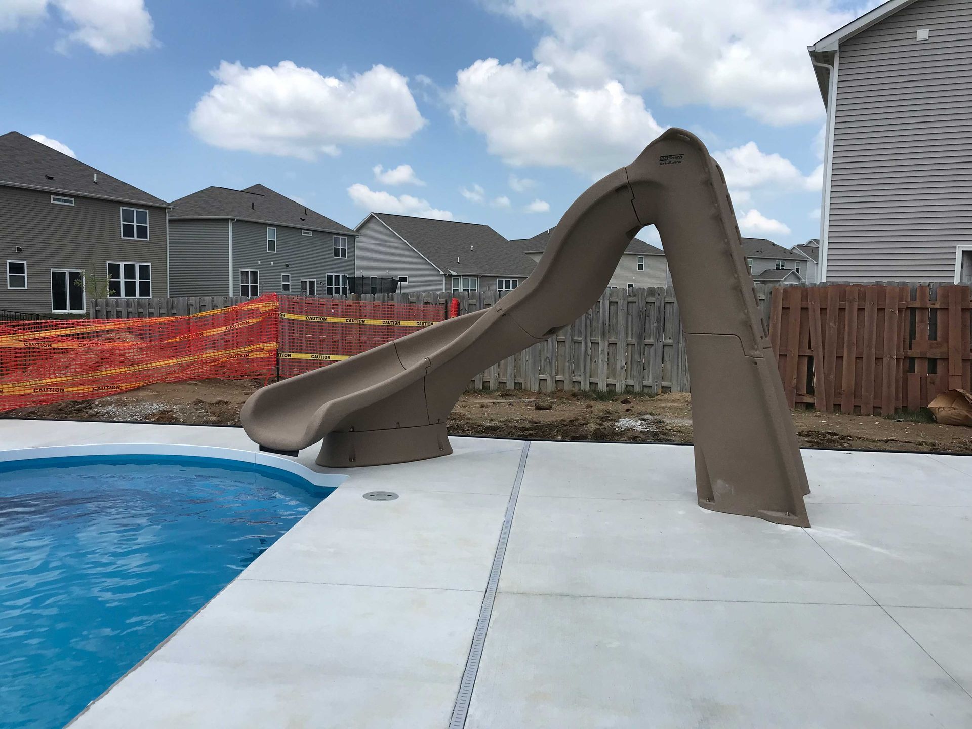 Brown pool slide on concrete next to a pool, houses in background.