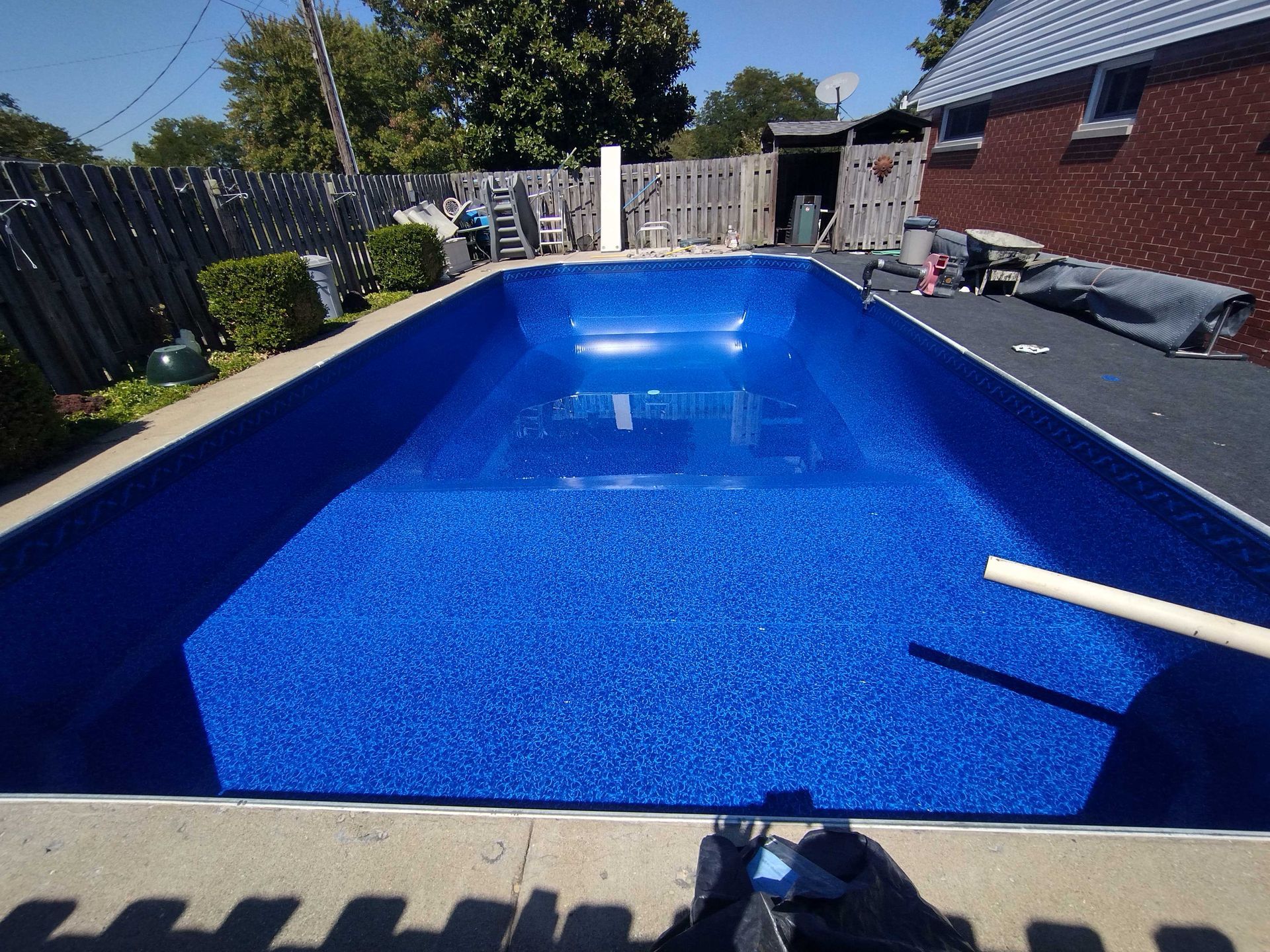 Blue-tiled rectangular swimming pool in backyard setting; wooden fence, brick house, and trees are visible.