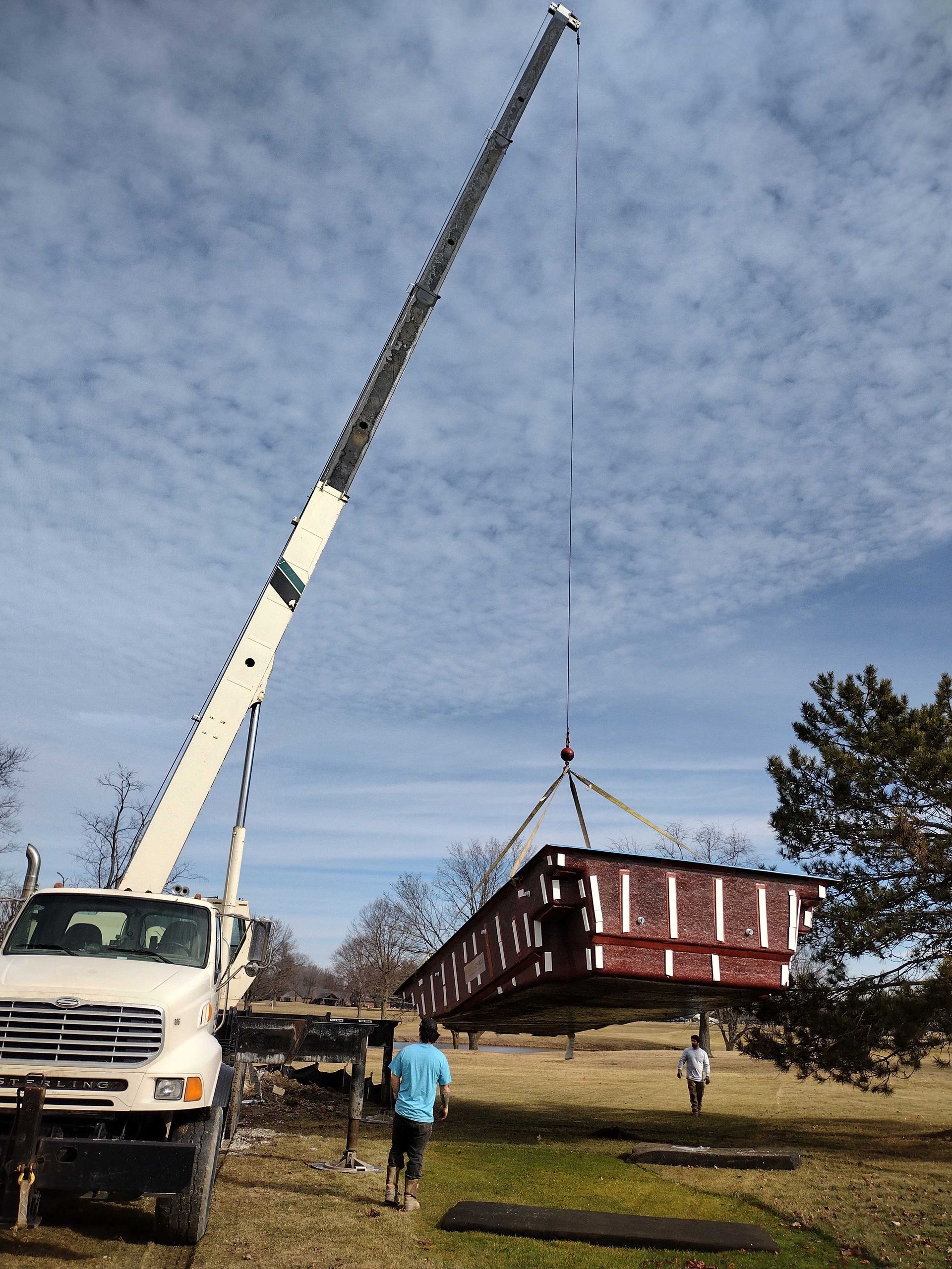 A crane lifting a large, rectangular red and brown object in a grassy field; two people stand nearby.