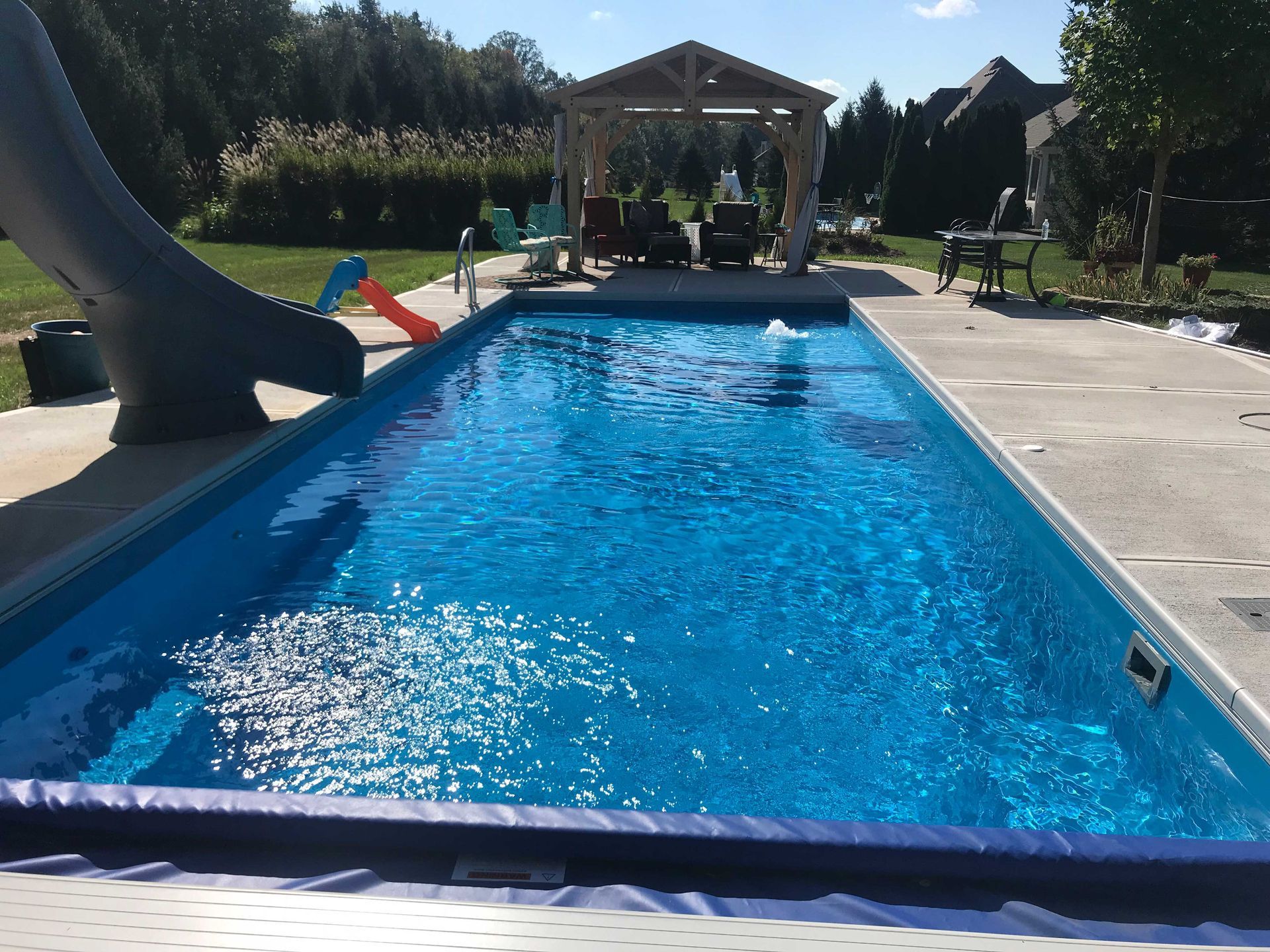 Rectangular blue pool with slide and gazebo in sunny backyard.