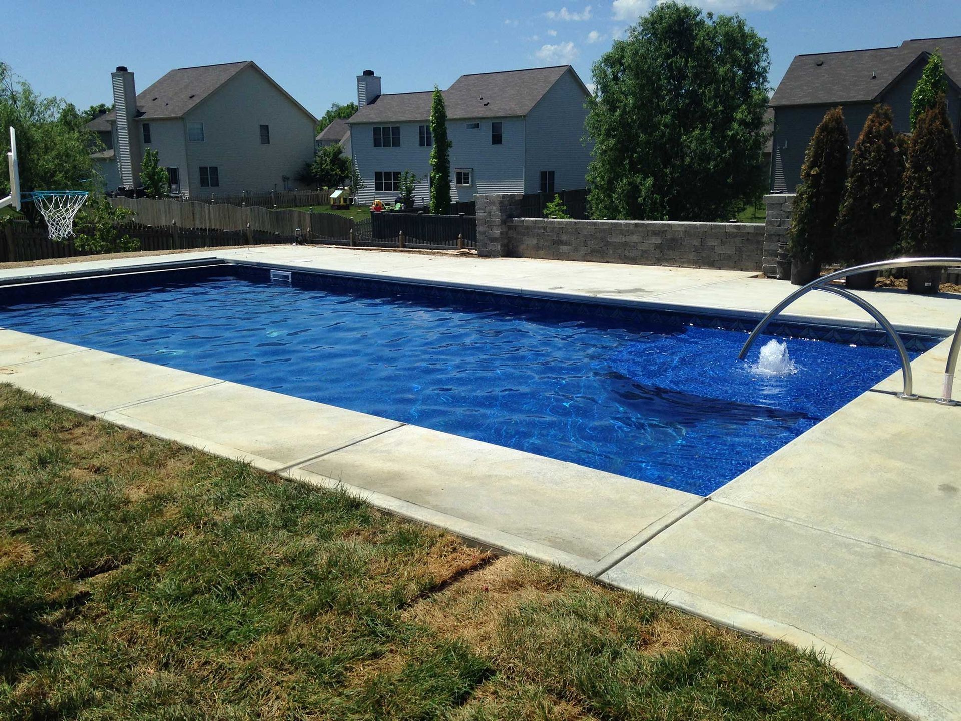 A backyard pool with blue water and concrete border, surrounded by grass, with houses in the background.