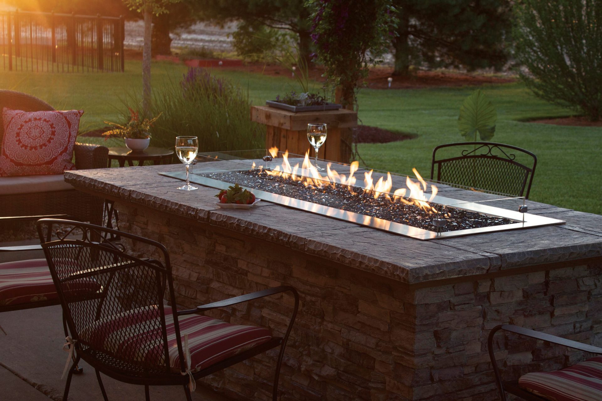 Backyard fire pit table glowing at dusk, with chairs and a wine glass on a patio.
